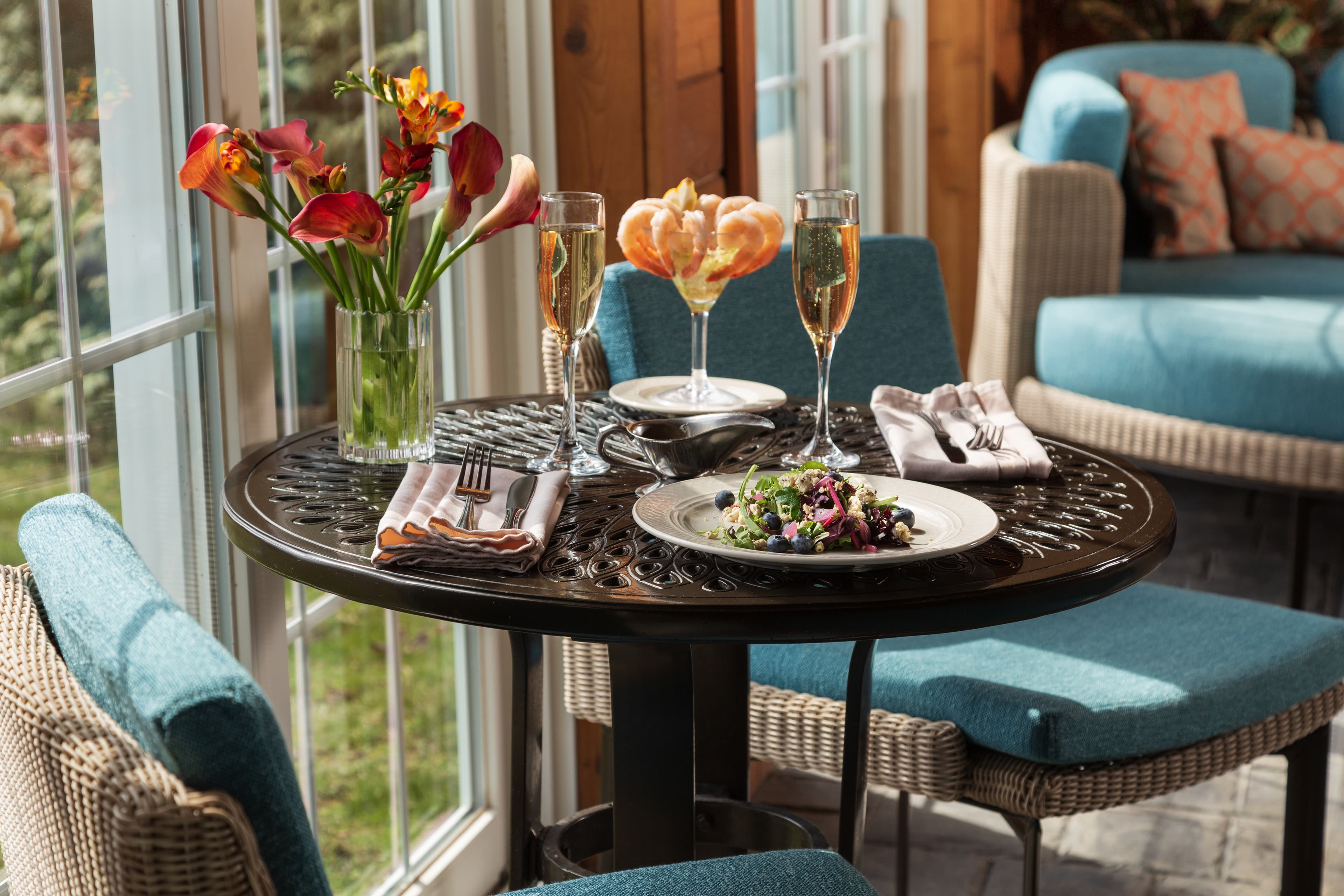 A cozy indoor seating area with large windows overlooking a lush green landscape. A round table with chairs is set for two, featuring a plate of food, champagne glasses, and flowers. The walls are paneled with wood, and there is a comfortable sofa with pillows in the background.