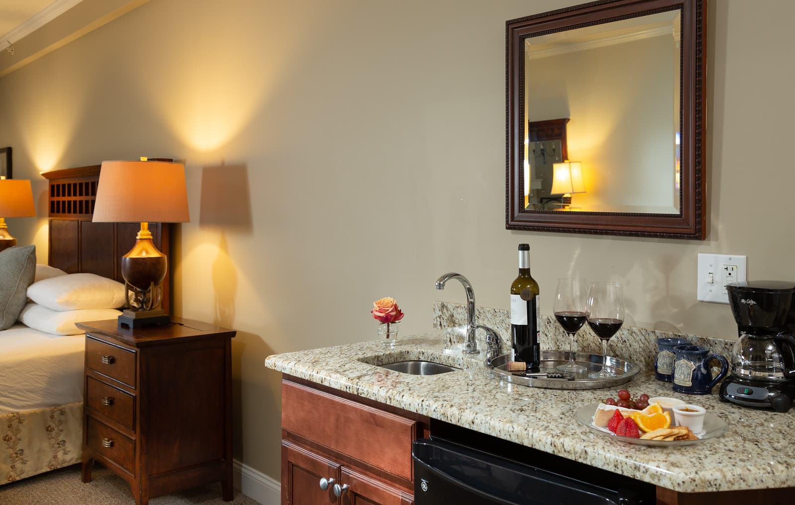 A cozy hotel room featuring a bedside table, a kitchenette with wine, glasses, and a plate of fruit.