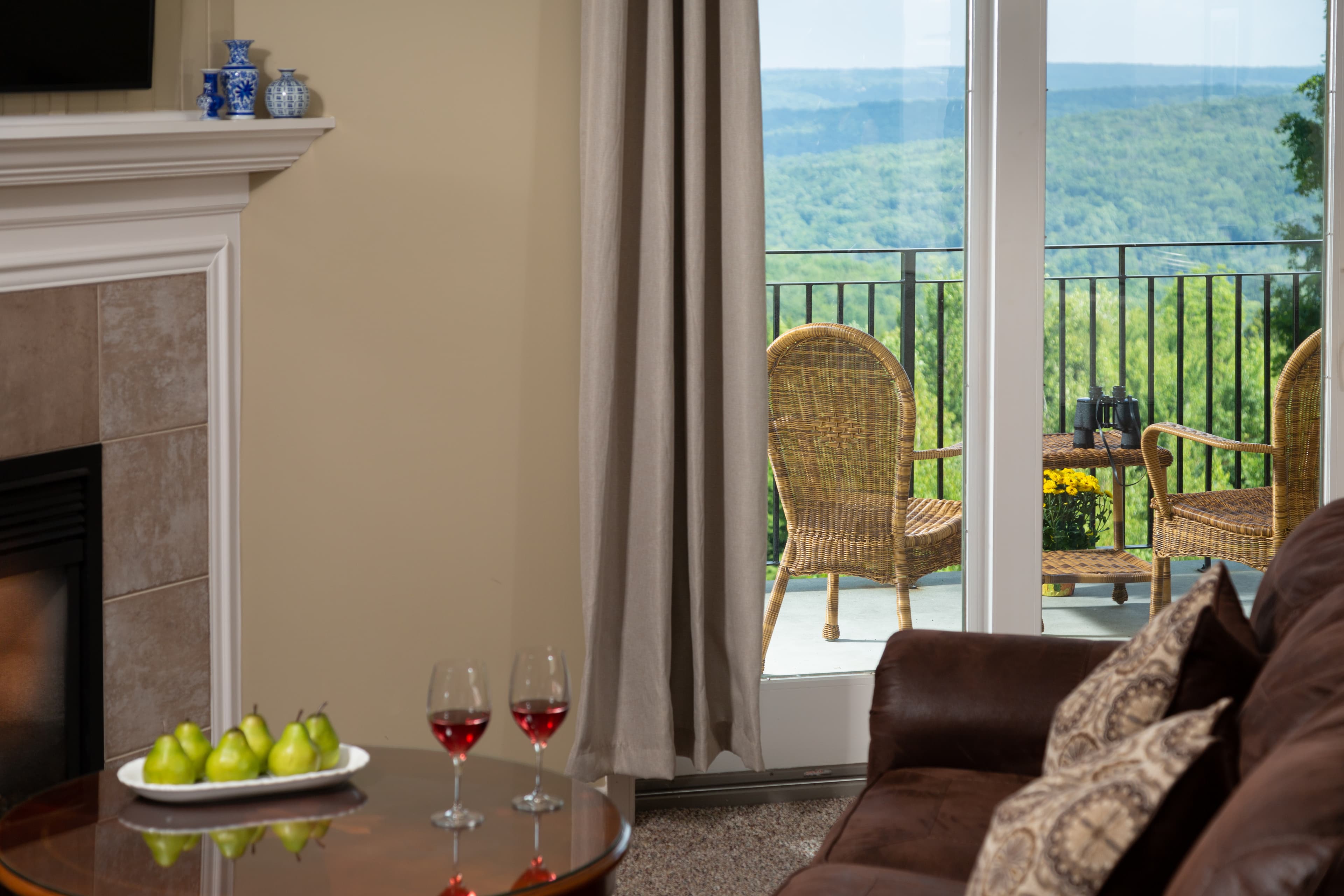 A cozy living area with a couch, a coffee table featuring pears and two glasses of red wine, and a view of a balcony overlooking a green landscape.