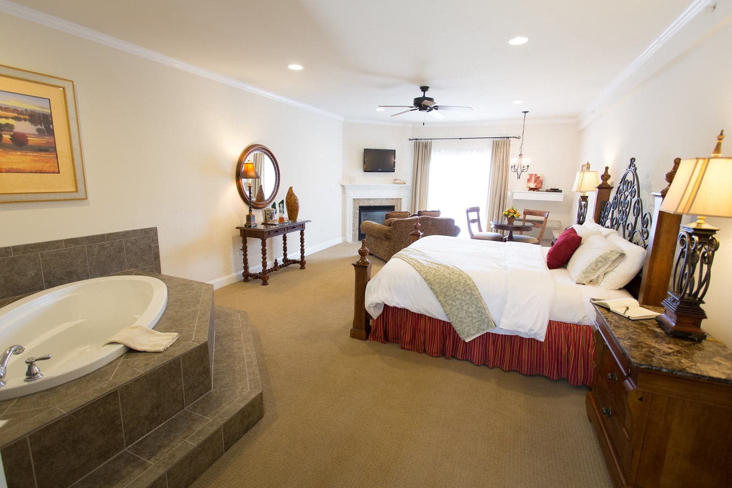 A spacious hotel room featuring a bed, armchair, large hot tub, and a small dining area.