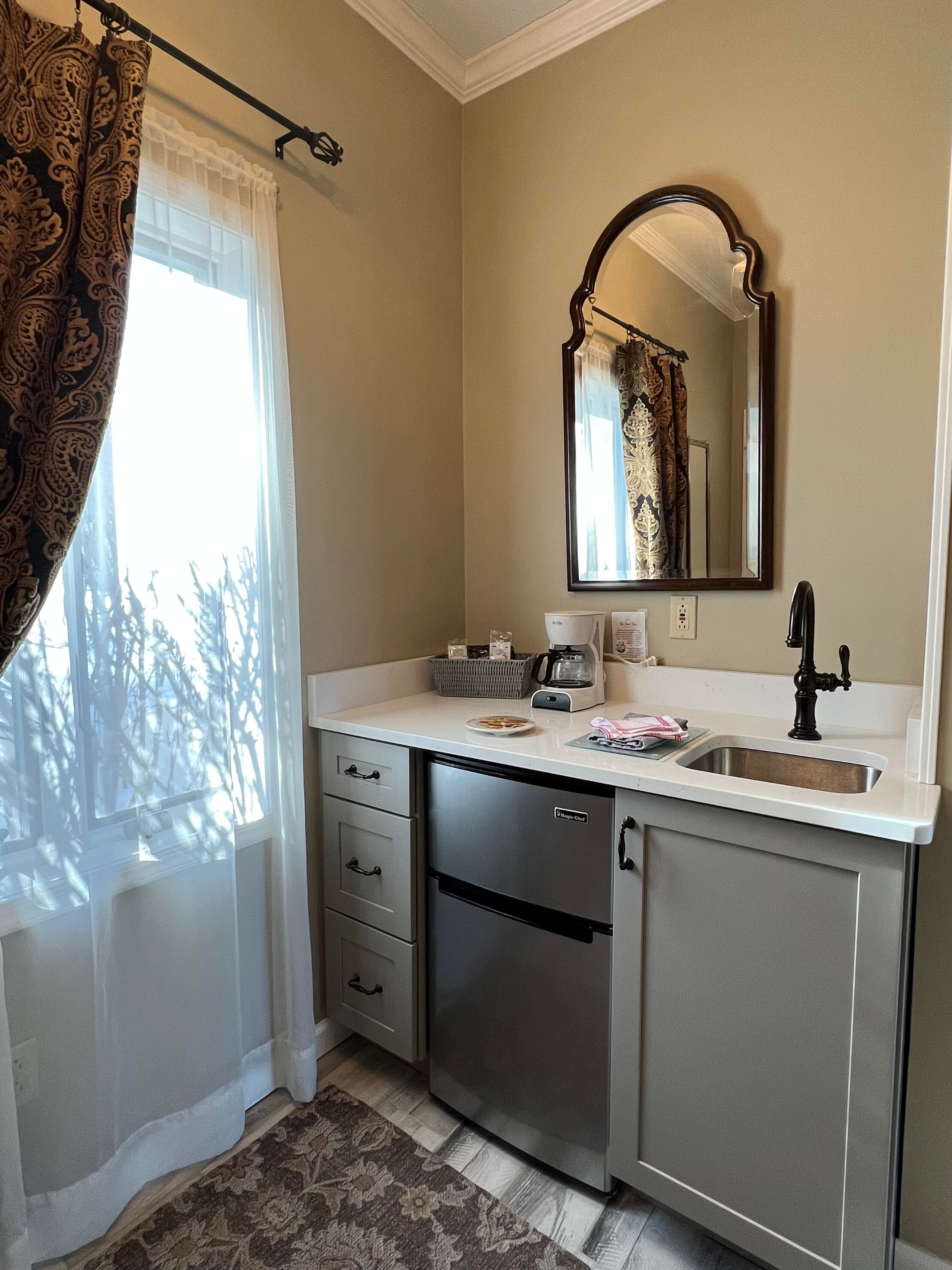 A small kitchenette featuring a stainless steel fridge, sink, and a mirror, with natural light from a nearby window.