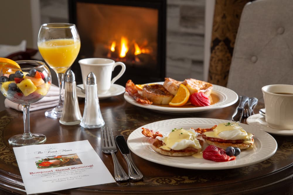 A delicious breakfast spread on a table in front of a fireplace. The meal includes French toast, eggs Benedict, fruit salad, orange juice, coffee, and bacon. The table is set with a white tablecloth, silverware, and a menu with the words "Bon Jour! Good Morning!" written on it.