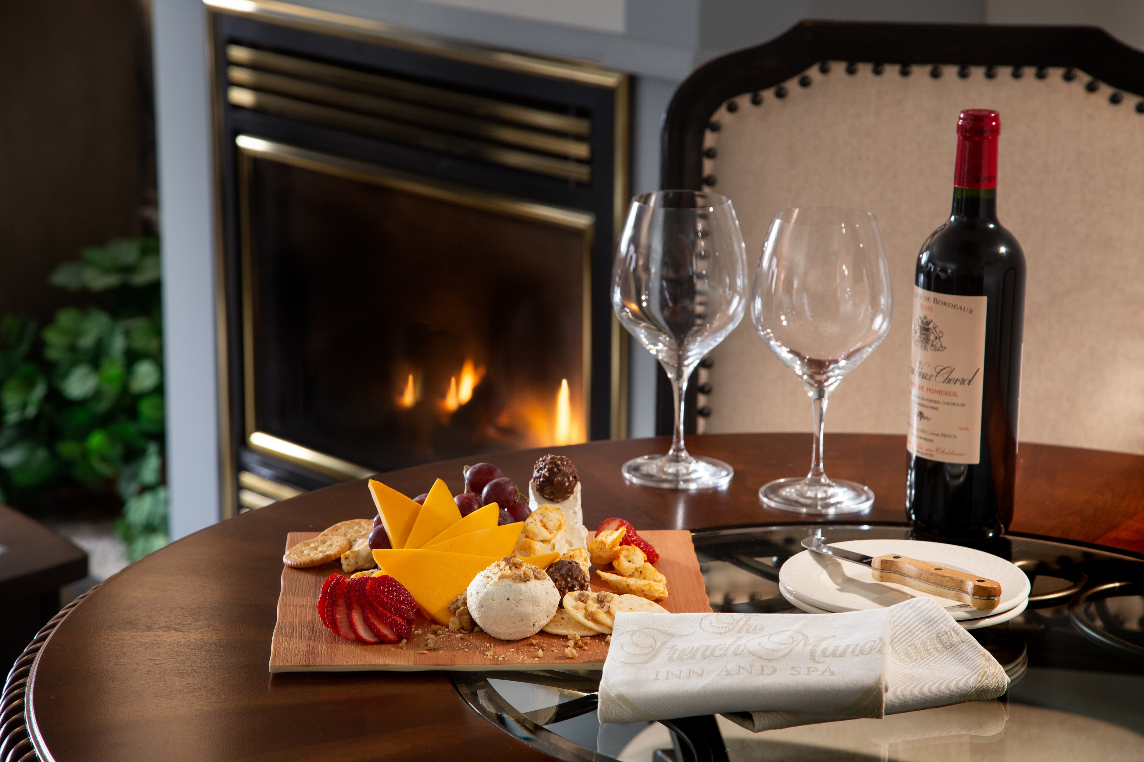 A cozy setup featuring a cheese platter, red wine, and glasses beside a warm fireplace.
