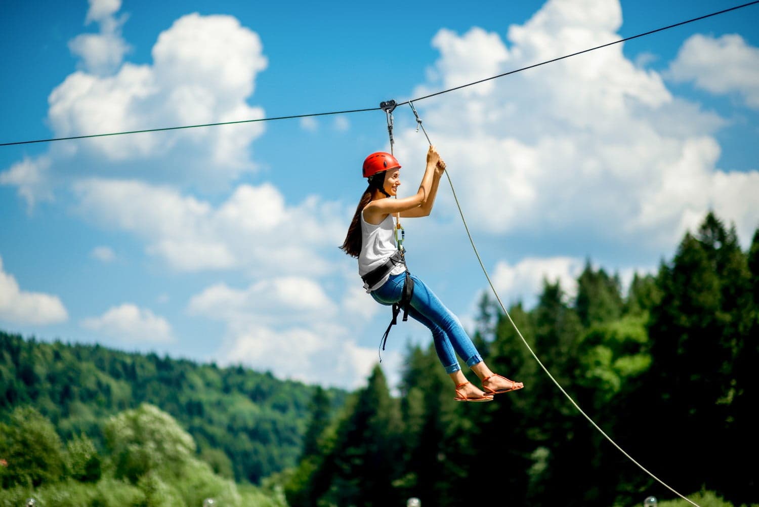 A young woman in a helmet zip lines through a scenic forested area under a bright blue sky.
