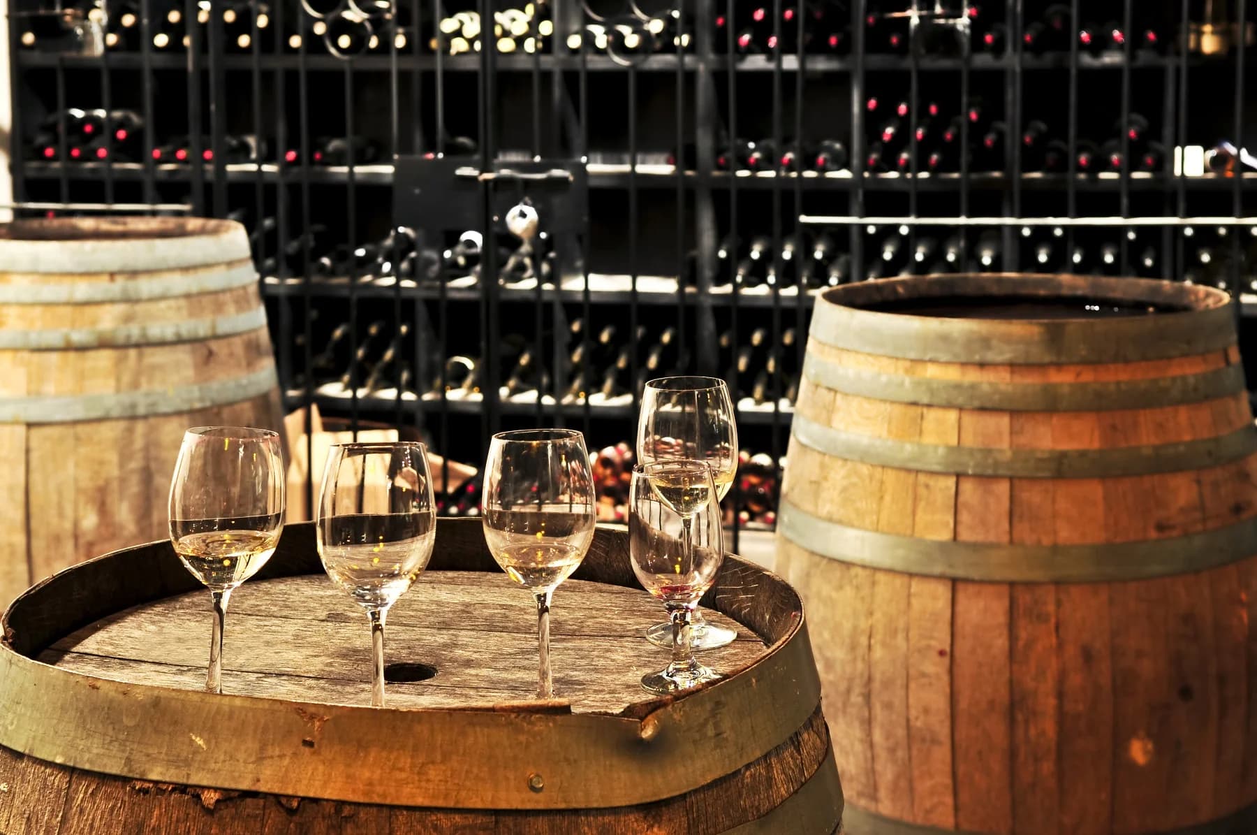 Four wine glasses are arranged on a wooden barrel in front of a wine rack filled with bottles.