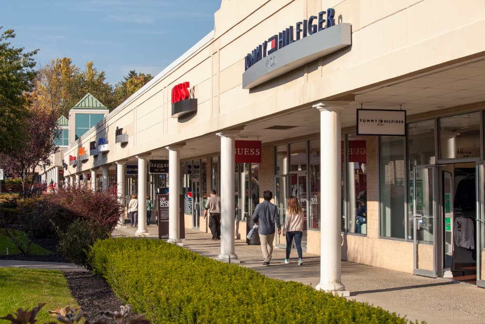 a row of shops such as Tommy Hilfiger and Guess with people walking along the sidewalk in front of the shops.