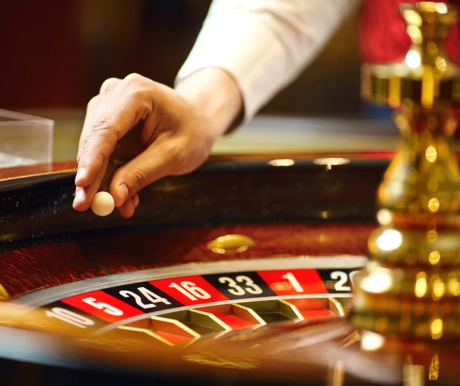 A hand poised to place a white ball on a roulette wheel at a casino.