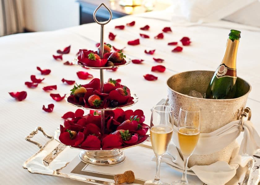 A silver tray holds a tiered stand of chocolate-covered strawberries, champagne flutes, and a bottle of champagne, surrounded by scattered rose petals. A silver tray holds a tiered stand of chocolate-covered strawberries, champagne flutes, and a bottle of champagne, surrounded by scattered rose petals.
