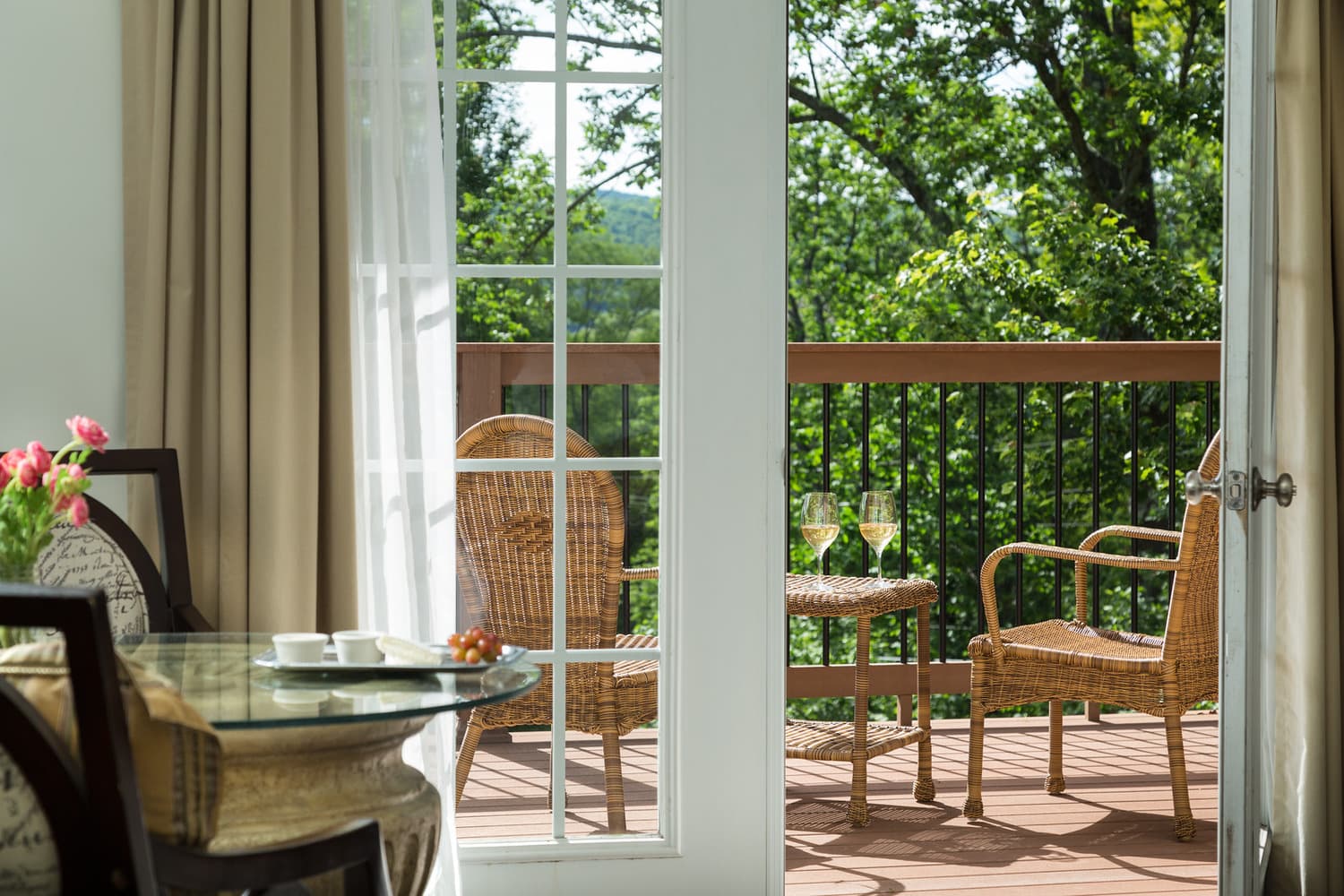 A view from inside a room through sliding glass doors onto a balcony with two chairs and a table, surrounded by greenery.