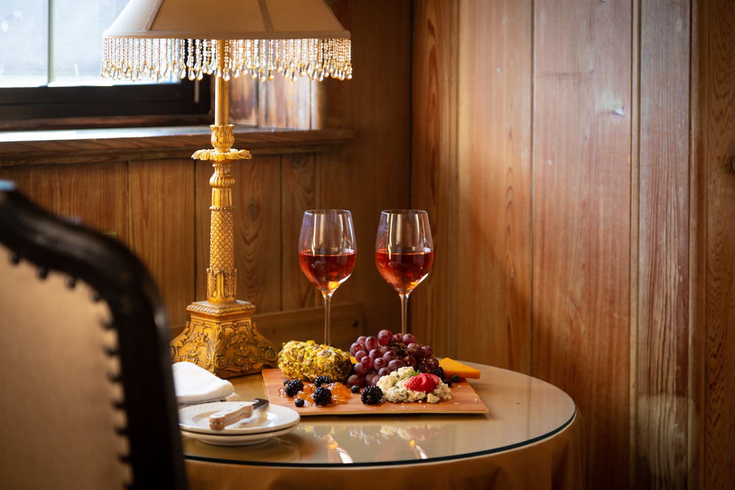 A cozy table setup featuring two glasses of rosé wine, a charcuterie board, and a decorative lamp.