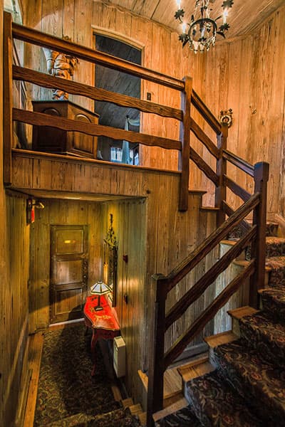 A wooden staircase leads to a warmly lit landing with a vintage lamp and an ornate door.