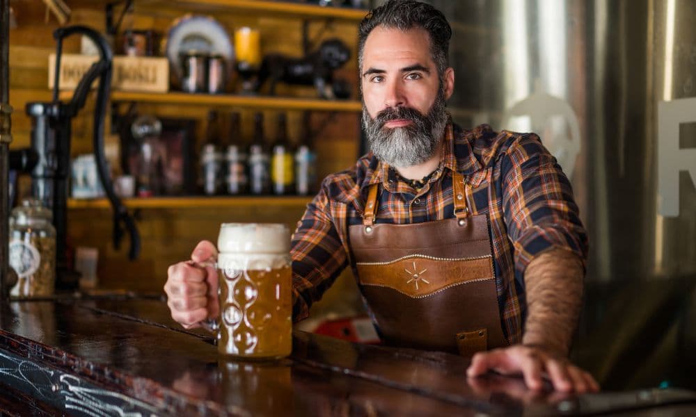 A bartender holding a glass of beer at a brewery bar, with brewing equipment and shelves in the background. A bartender holding a glass of beer at a brewery bar, with brewing equipment and shelves in the background.