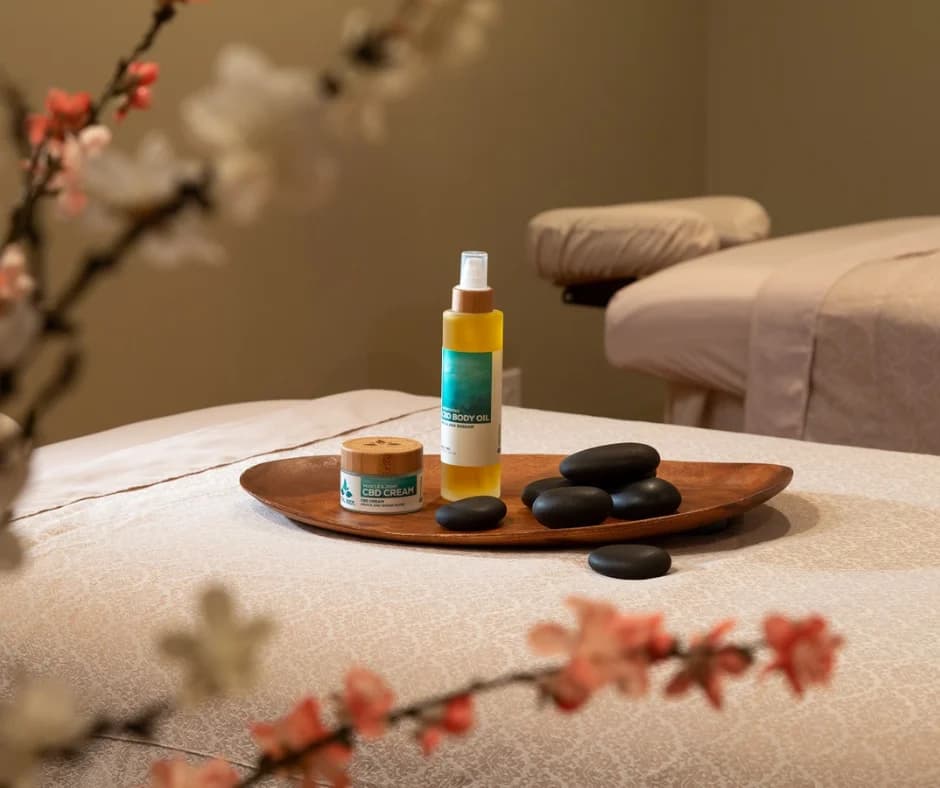 A wooden tray on a massage table holds stones, a bottle of oil, and a jar of cream, surrounded by delicate flowers in the background.