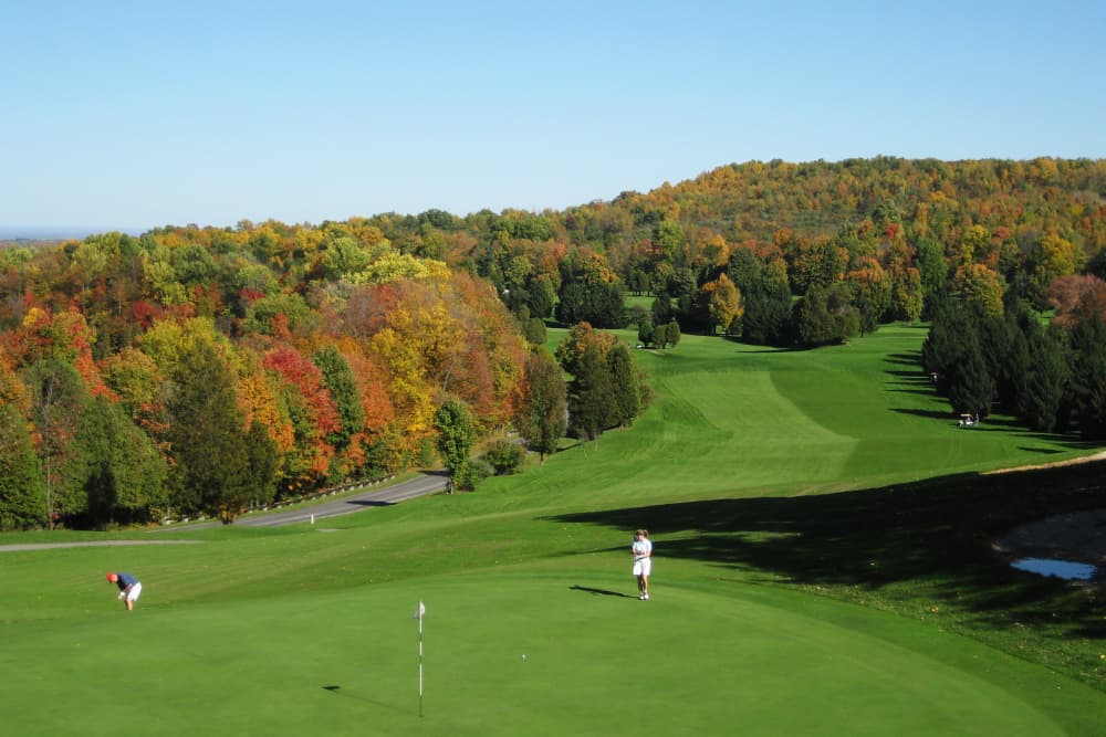 A scenic golf course with a golfer putting on a green. The course is surrounded by colorful autumn foliage and rolling hills.