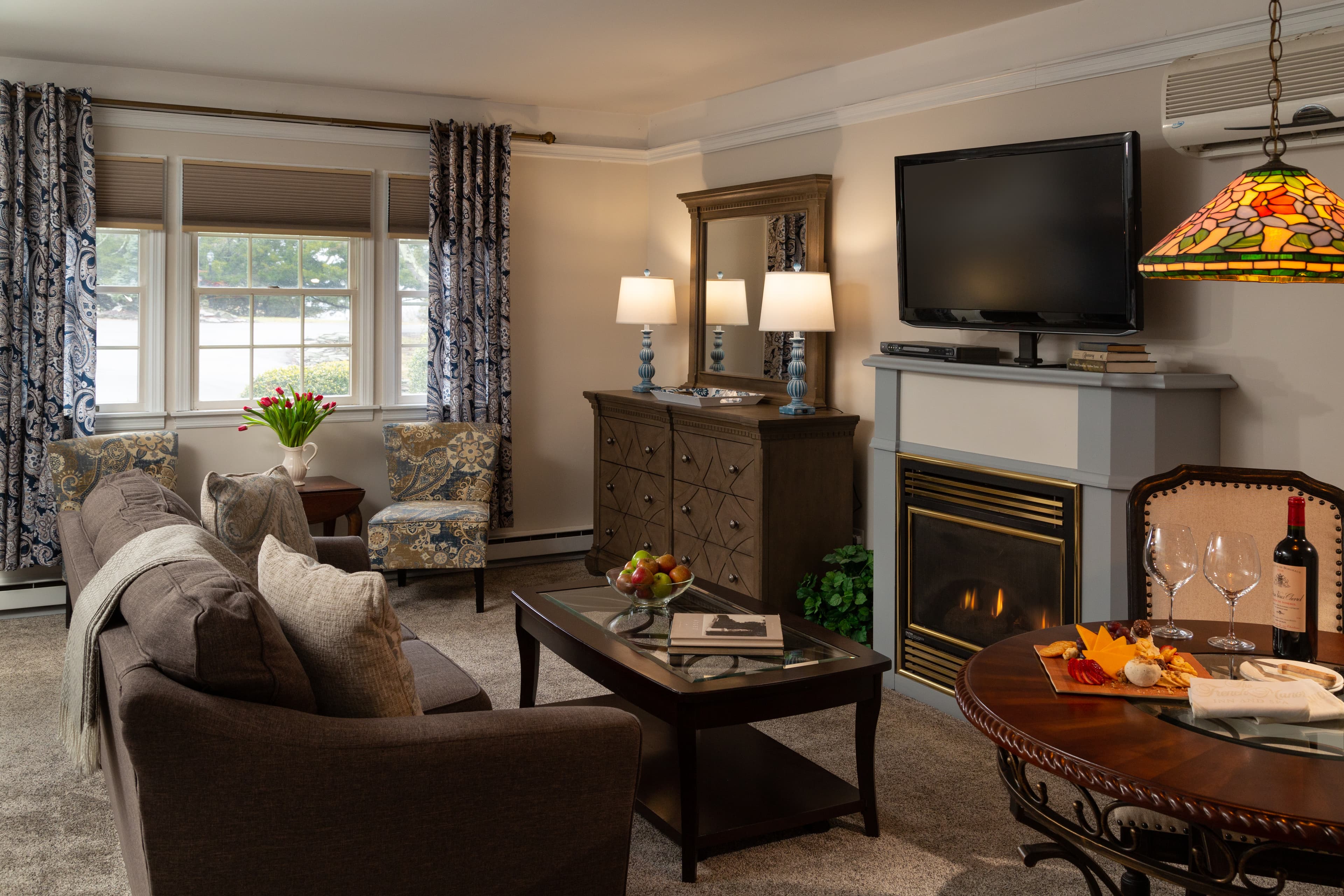 Cozy living room with a gray sofa, decorative chairs, a fireplace, and a coffee table set with wine and snacks.