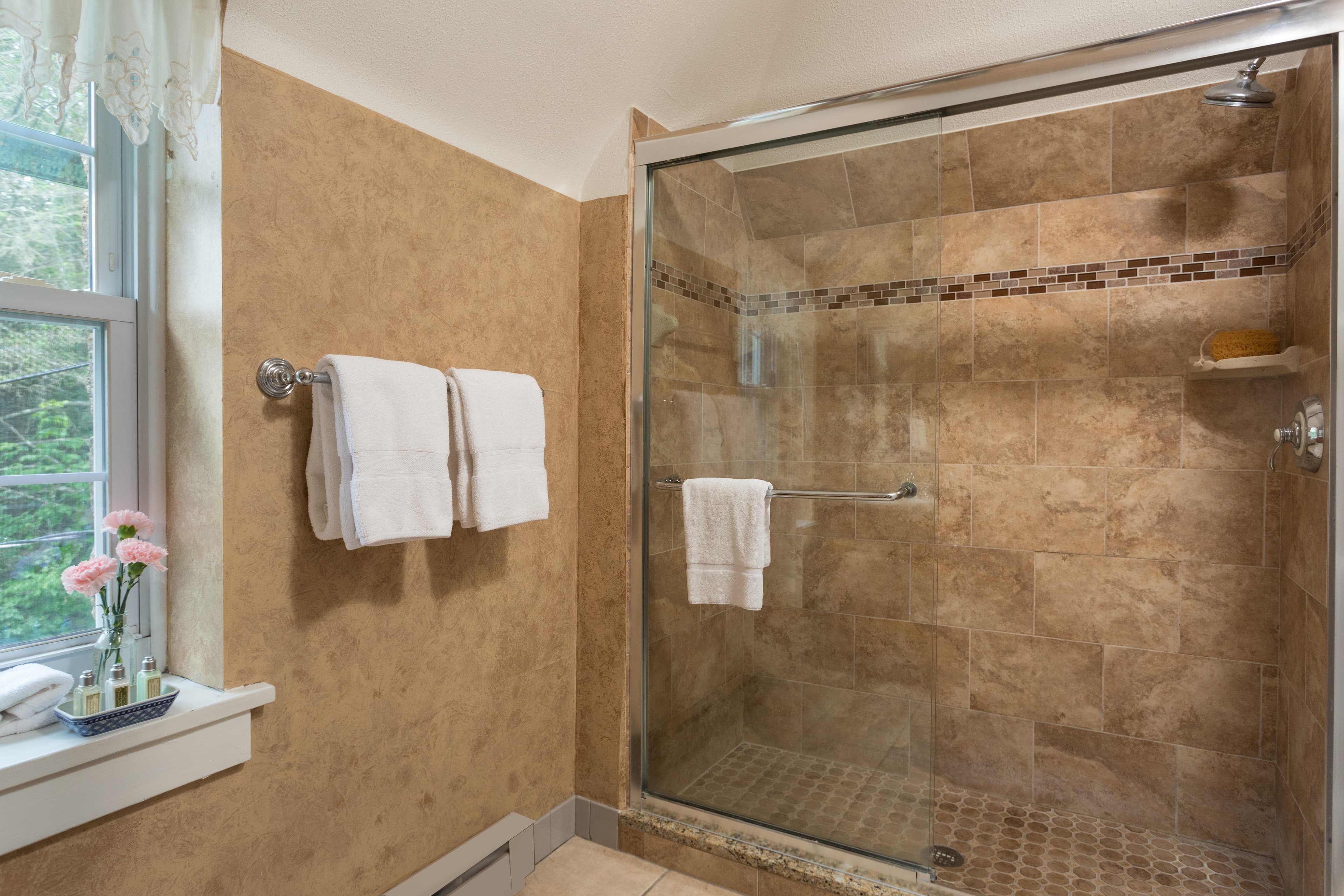 A modern glass shower with beige tile and fresh towels hanging nearby in a well-lit bathroom.