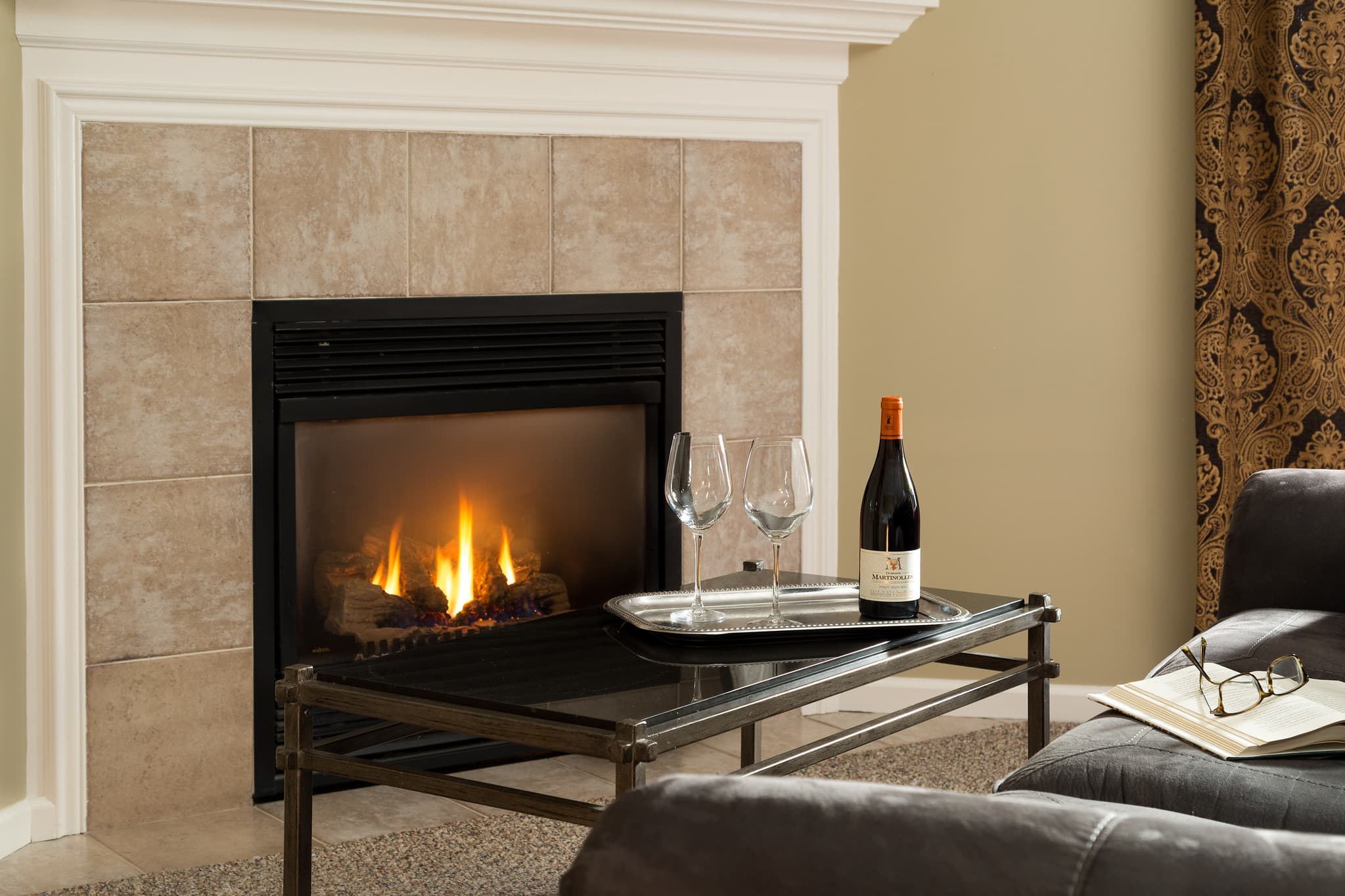 A cozy living room scene featuring a lit fireplace, a tray with wine glasses and a bottle, and a pair of glasses on a table.