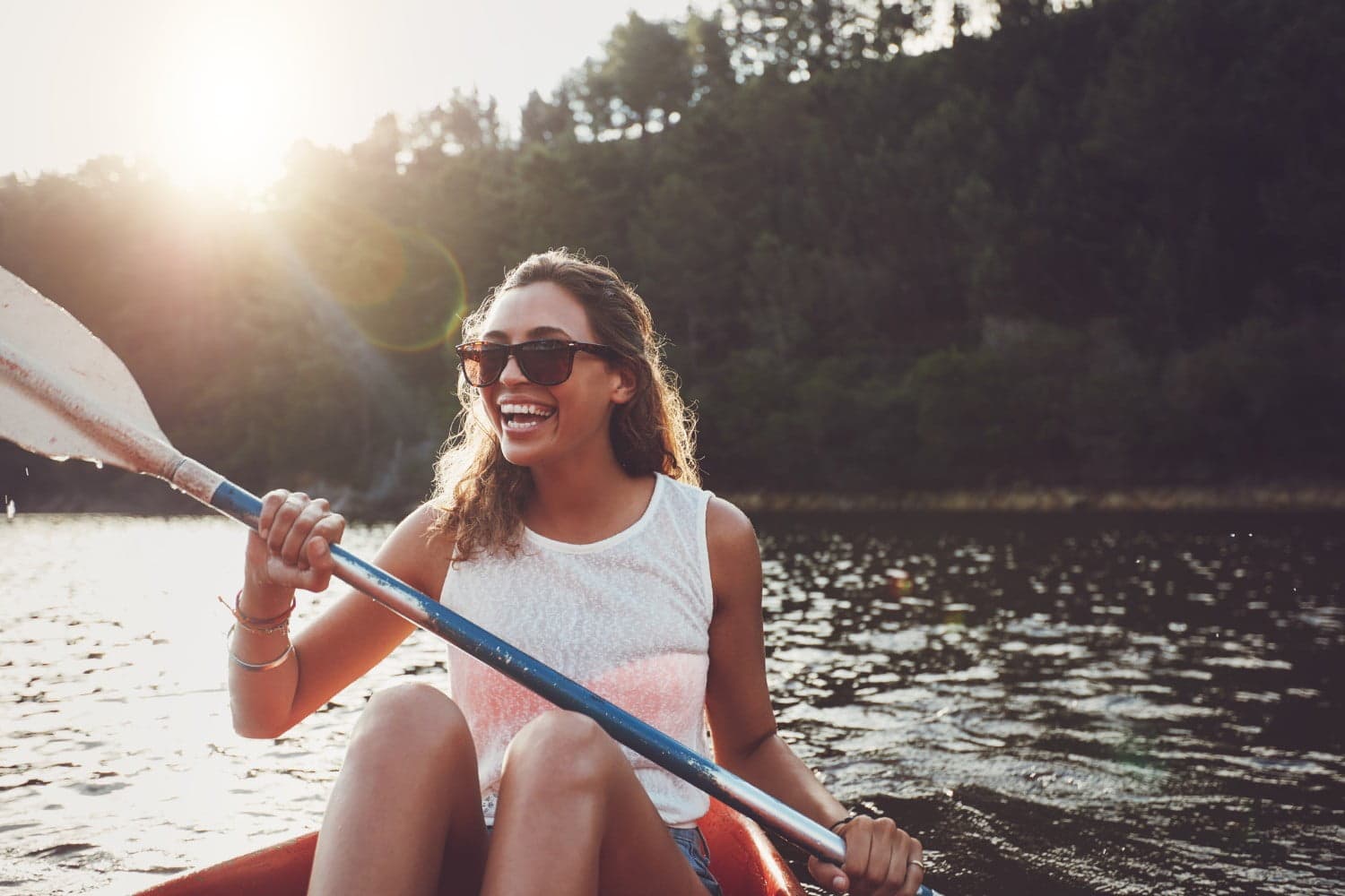 A woman with curly hair and sunglasses smiles while kayaking on a calm lake. A woman with curly hair and sunglasses smiles while kayaking on a calm lake.