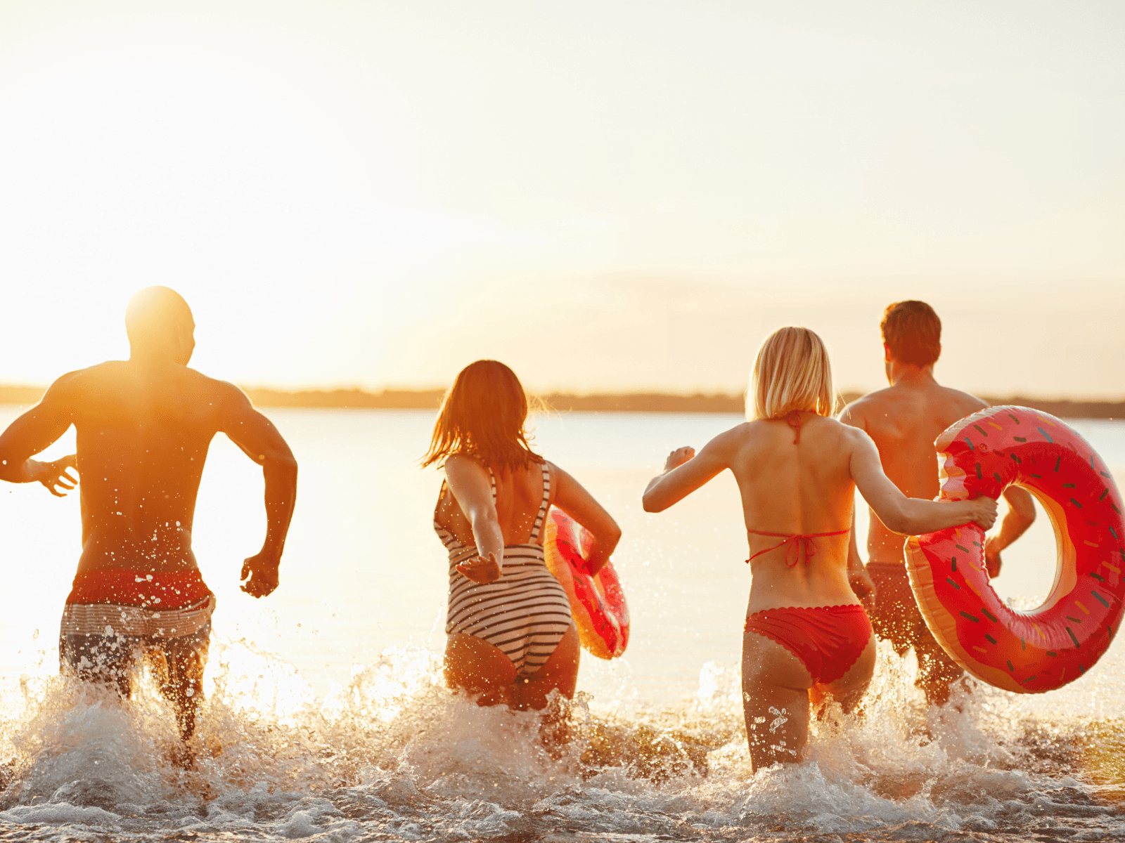 Four people wade into the water at sunset, holding colorful inflatable rings.