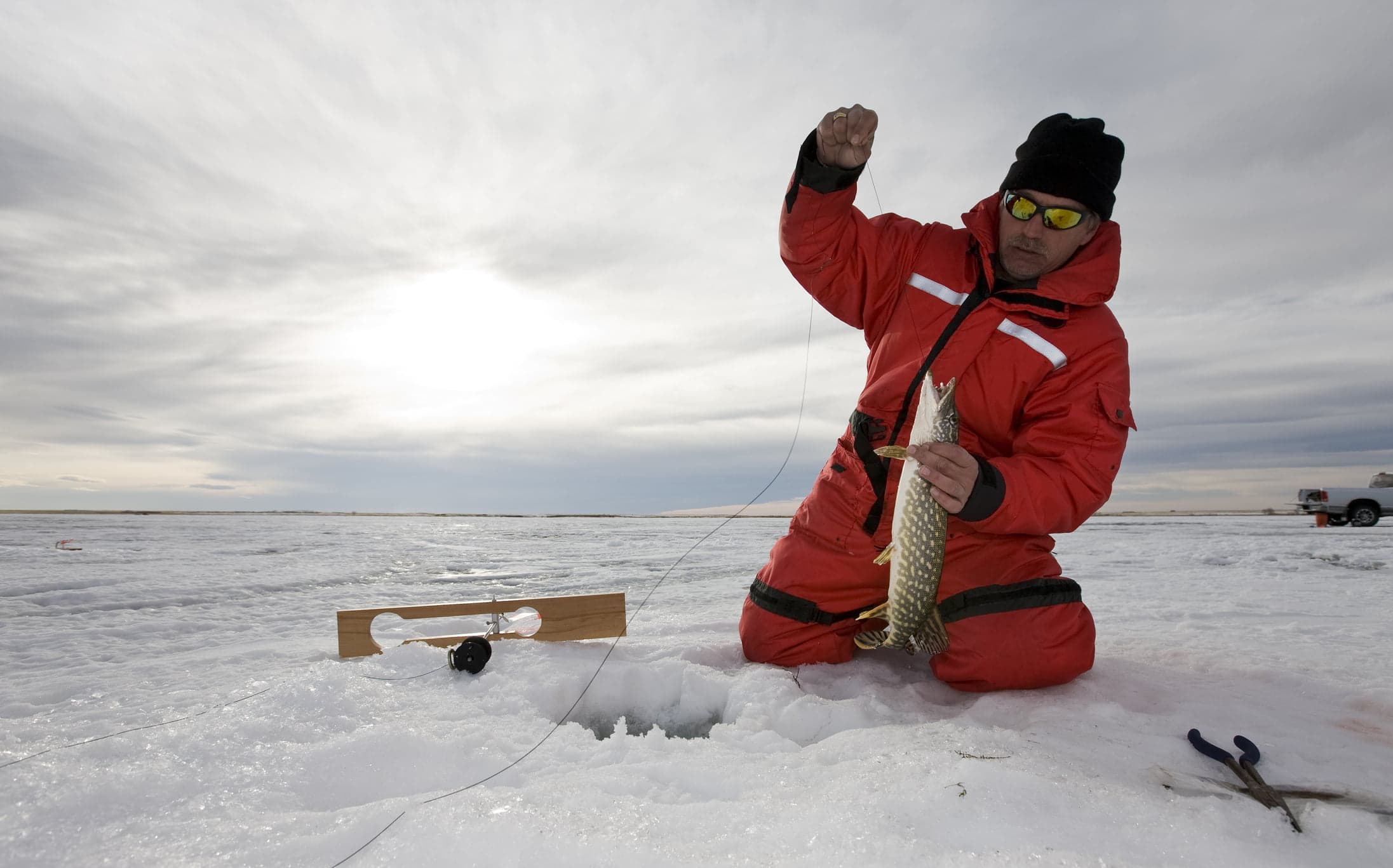 A fisherman in a red suit pulls a fish from a hole in the ice against a cloudy sky.