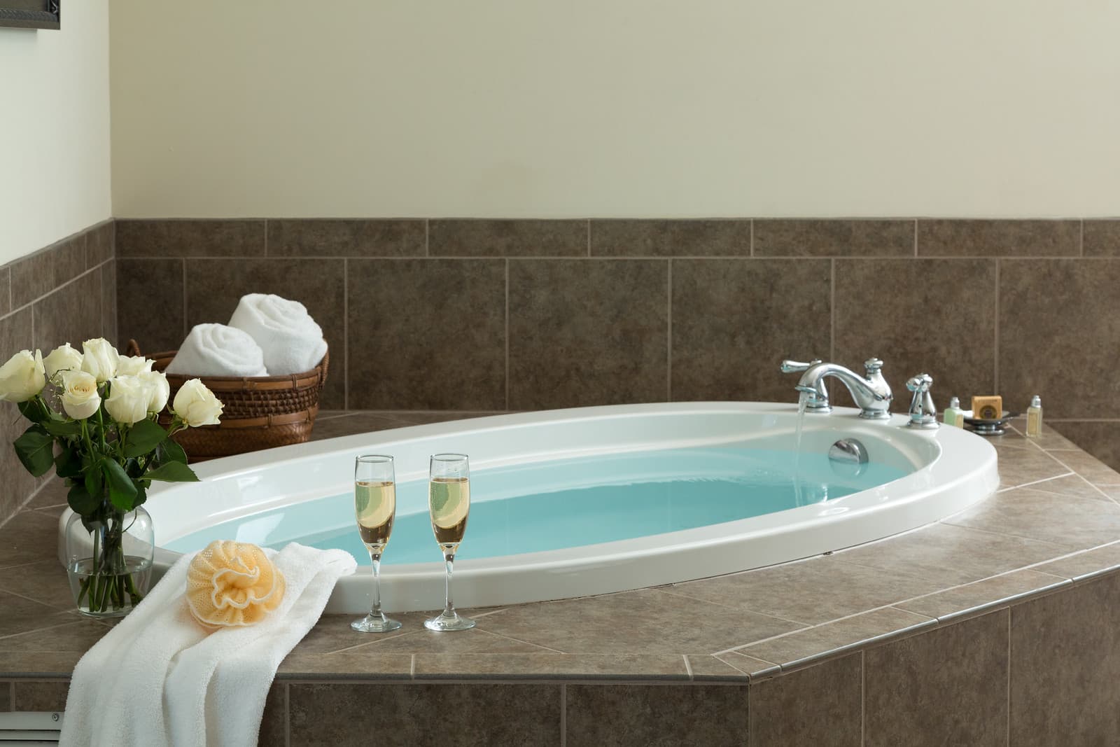 A luxurious bathtub with pale blue water, decorated with white roses, a towel, and two glasses of champagne.