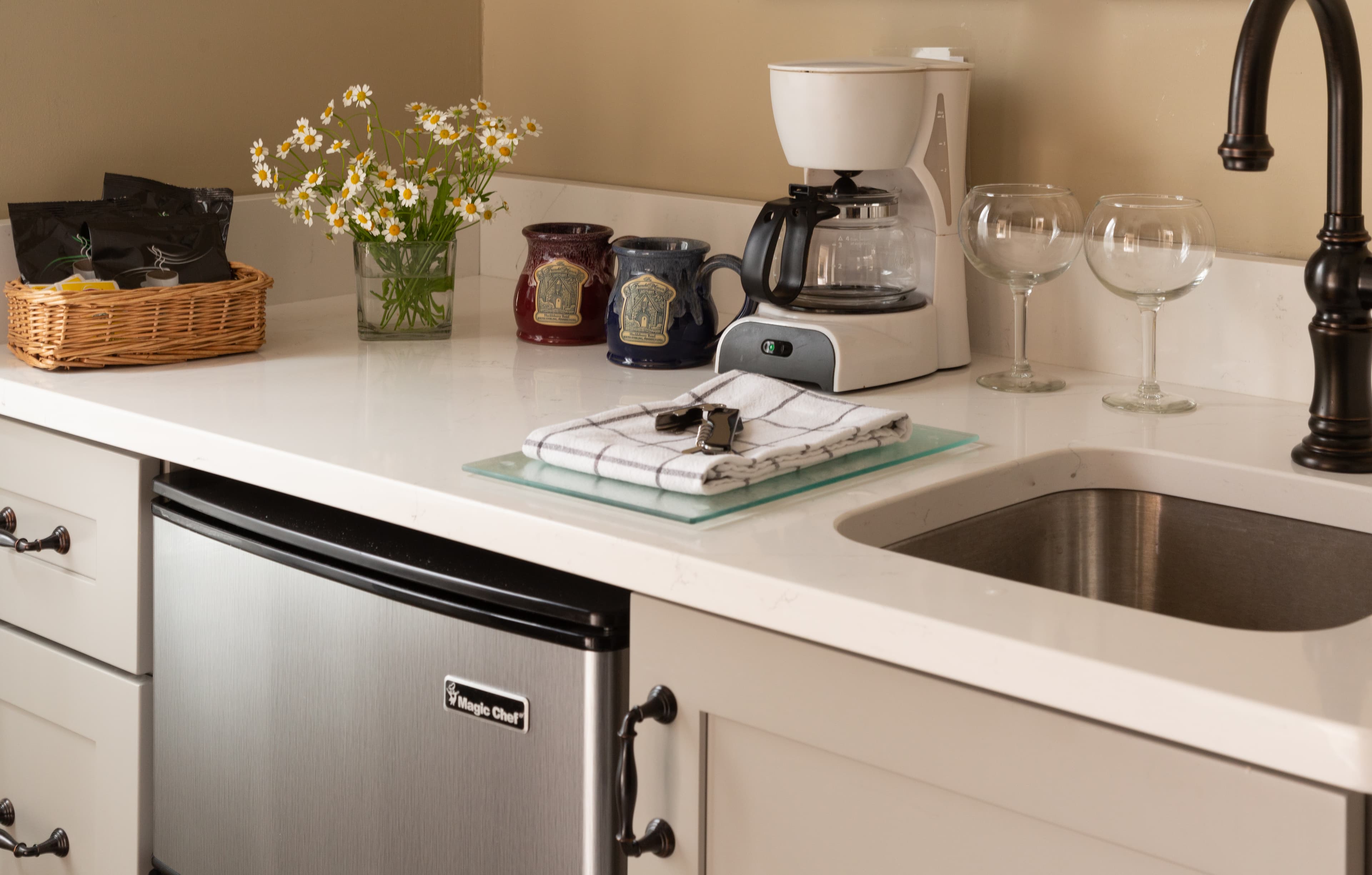 A kitchen countertop featuring a coffee maker, glasses, a small sink, and decorative flowers in a basket.