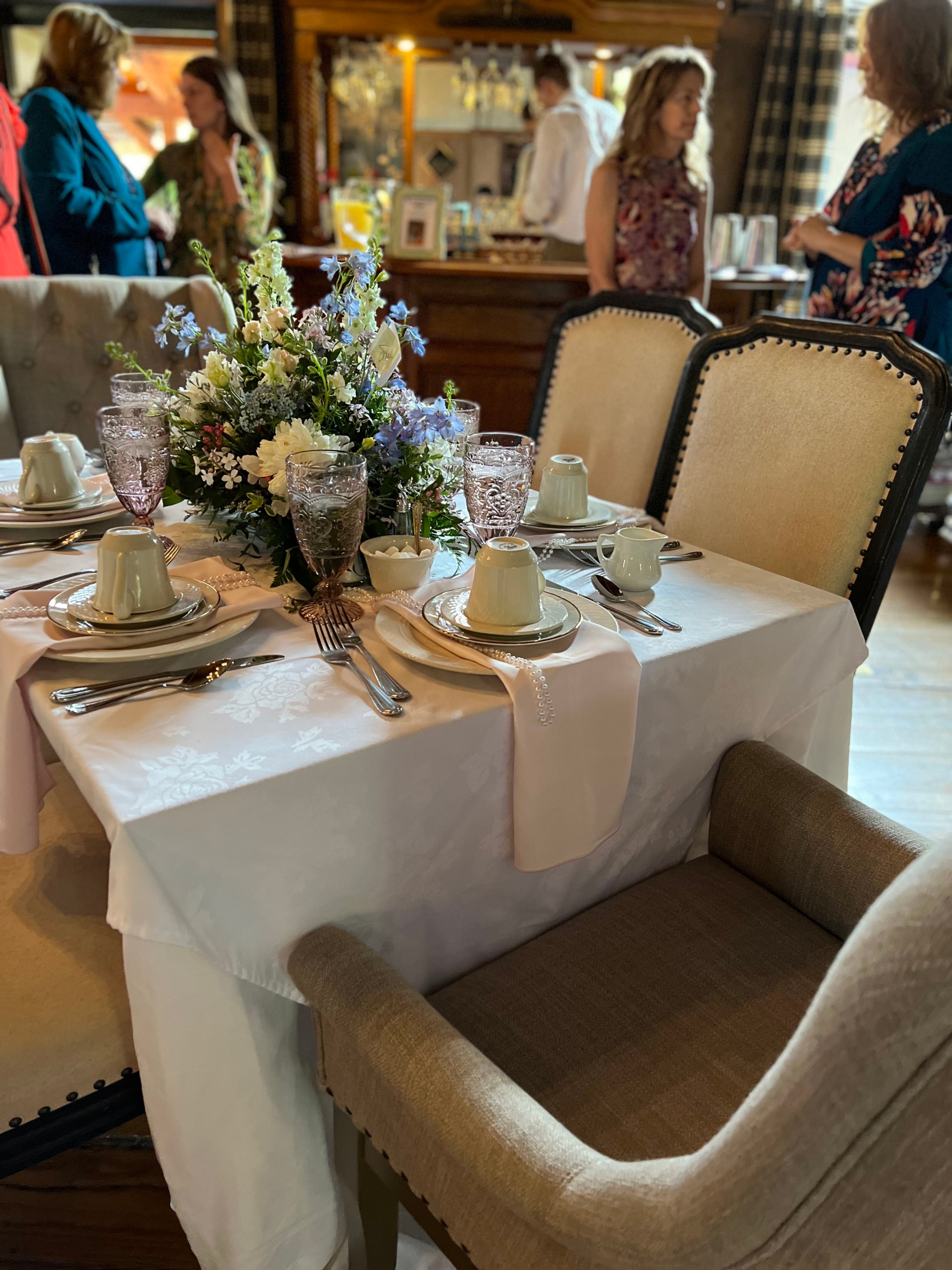 a group of people visiting in a room with a table elegantly set for a brunch or shower with a large flower centerpiece in the middle of the table.