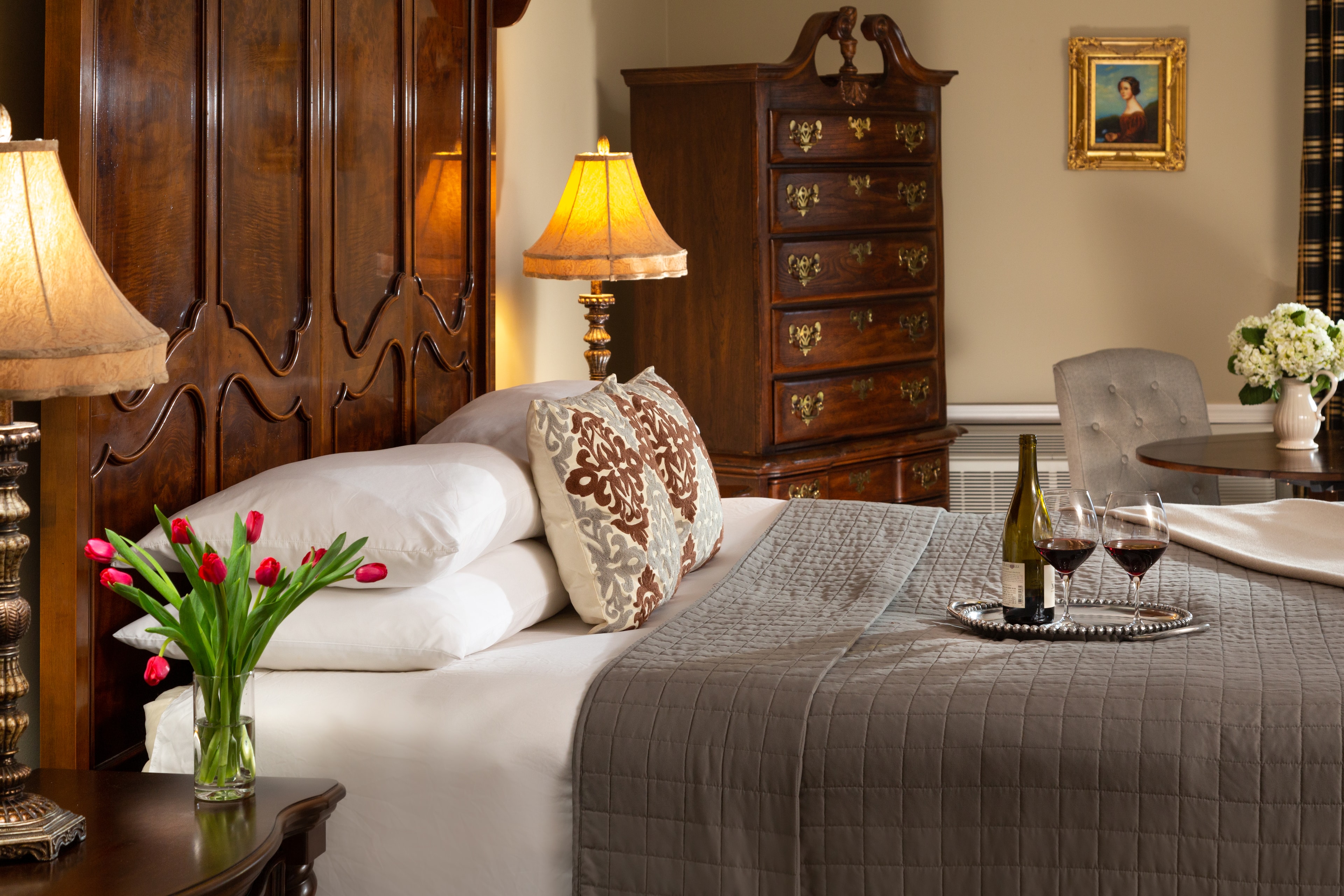 A cozy bedroom featuring a beautifully made bed, decorative pillows, bedside lamps, a vase of tulips, and a small table with wine glasses and a pitcher.