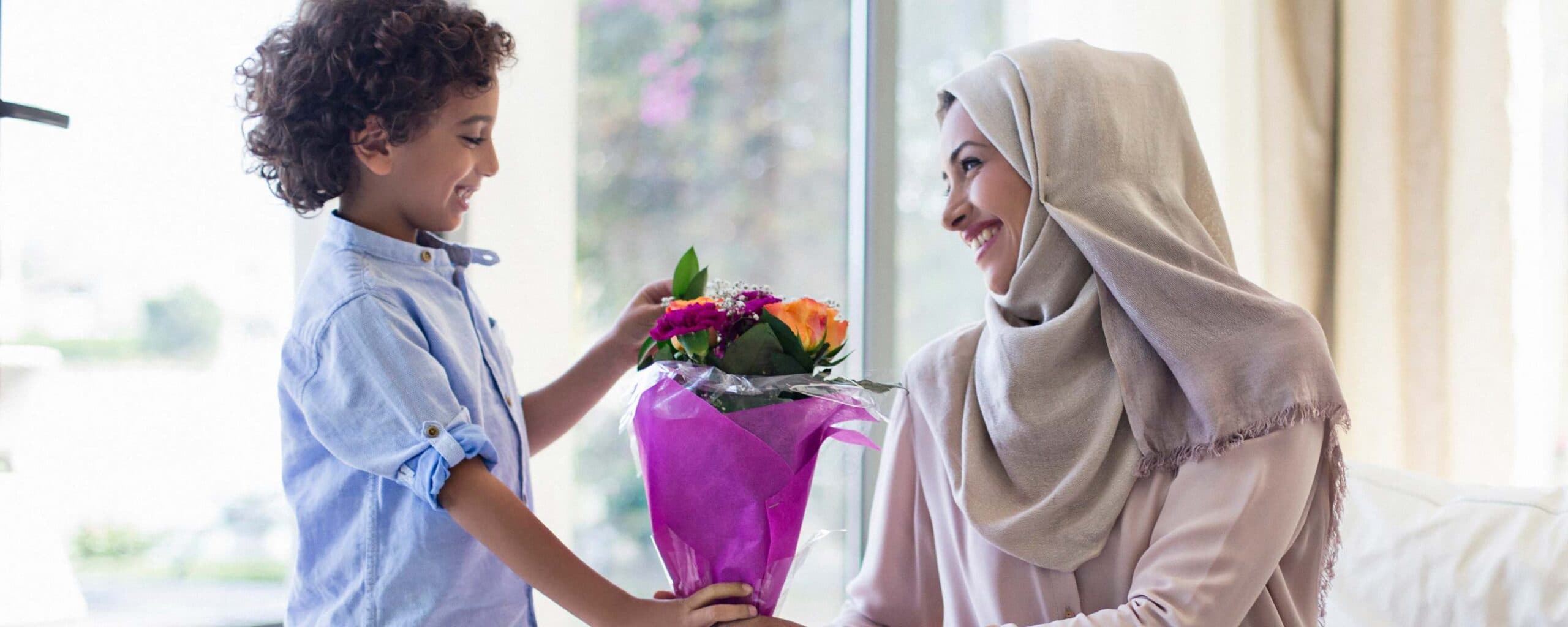 A young boy hands a bouquet of flowers to a smiling woman wearing a hijab. A young boy hands a bouquet of flowers to a smiling woman wearing a hijab.