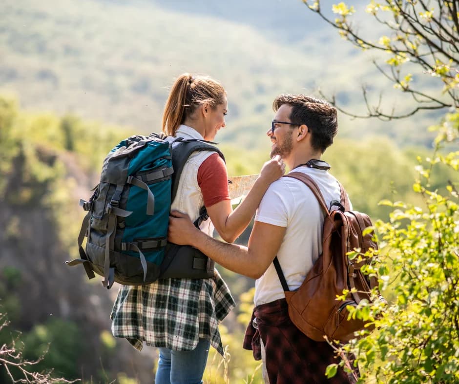 A couple with backpacks smiles at each other while looking at a map in a scenic outdoor setting.