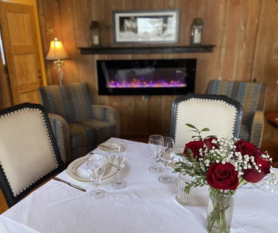 A elegantly set dining table with roses and wine glasses, featuring a cozy seating area and a modern fireplace in the background.