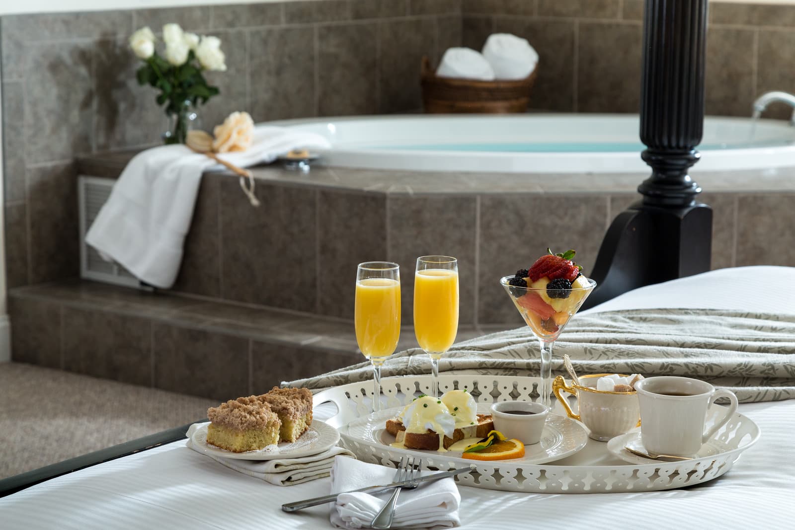 A luxurious hotel room featuring a tray of breakfast items with drinks, alongside a jetted tub and fresh flowers.