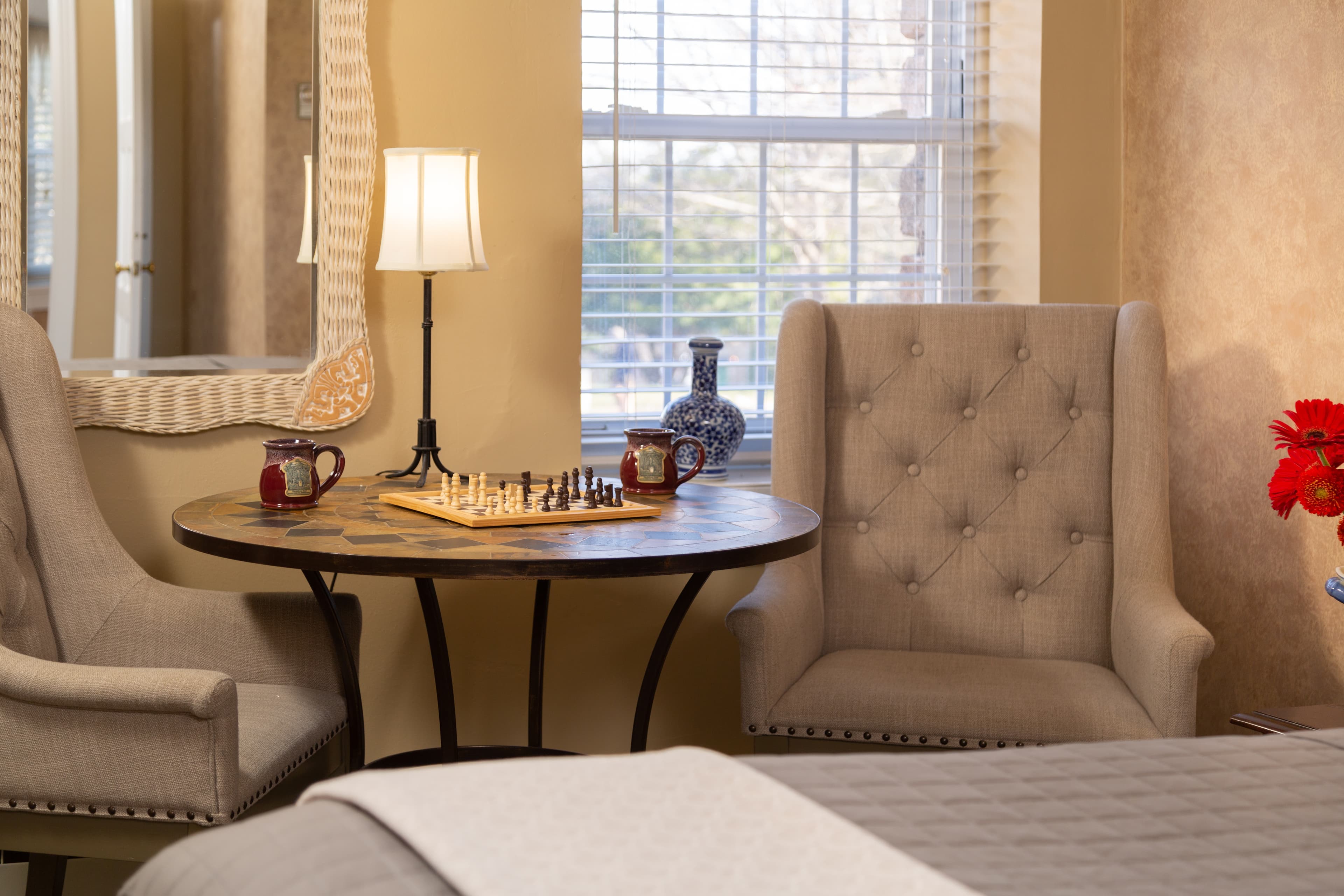 A cozy corner featuring two tufted armchairs, a small table with a chess set, and a lamp by a sunny window.