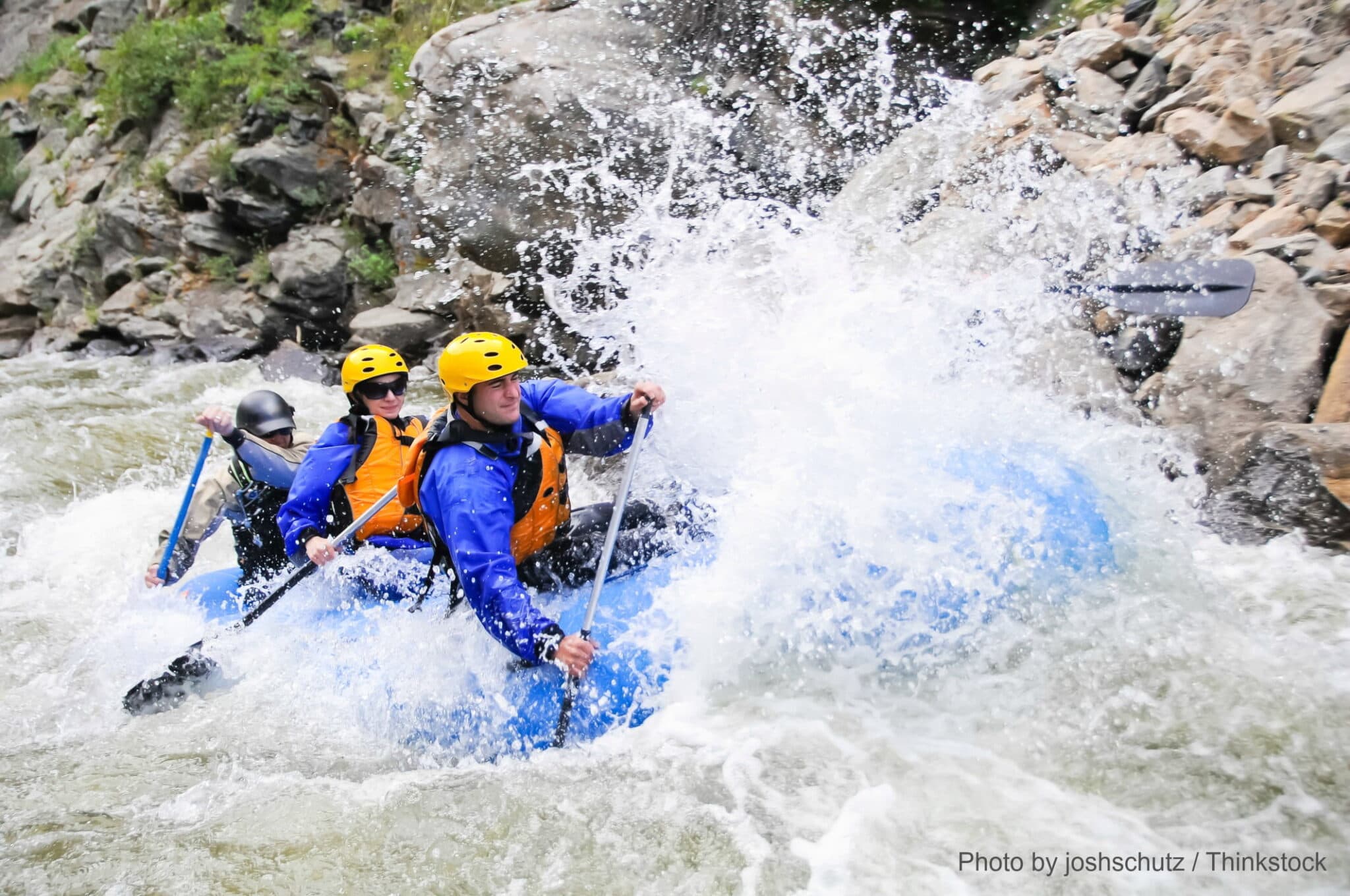 Three people in a blue raft navigate turbulent waters while wearing helmets and life jackets.