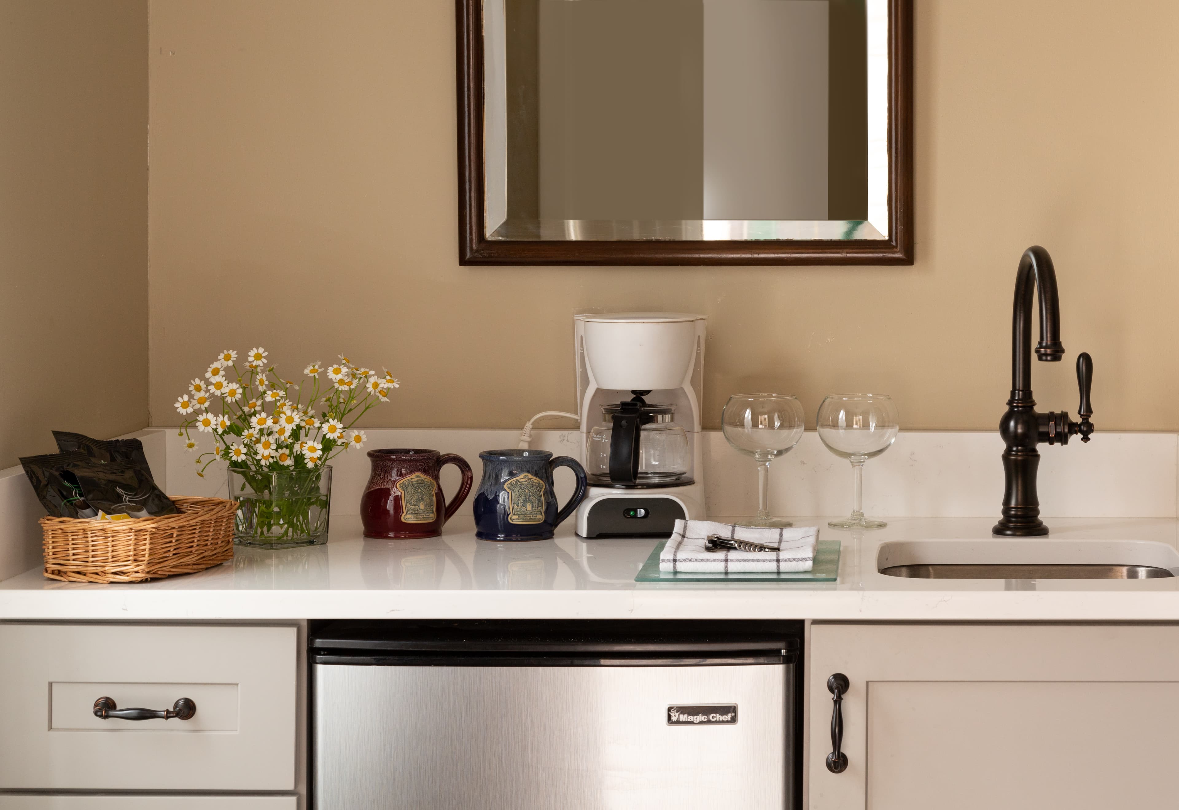 A stylish kitchen countertop featuring a coffee maker, glassware, flowers, and a decorative basket.