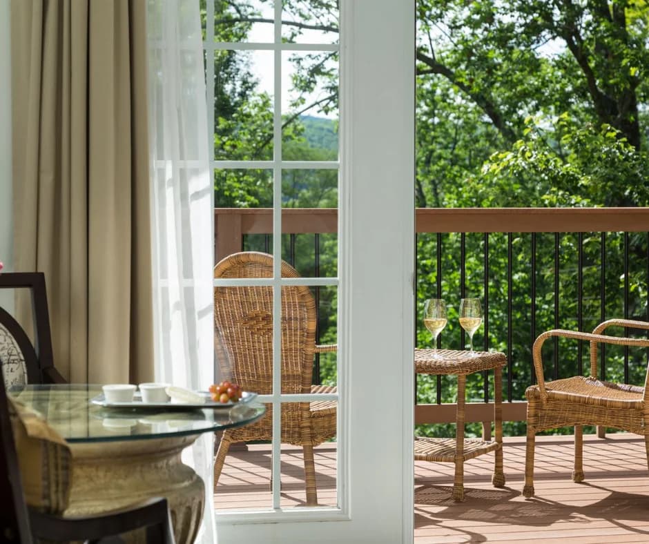 A bright interior view of a balcony with two chairs, a table with wine glasses, and greenery in the background.