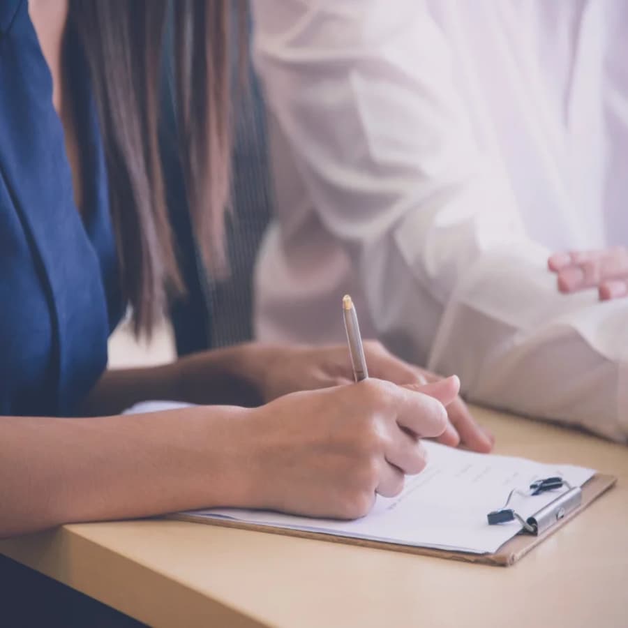 A man and a woman sitting at a table with a woman writing on a piece of paper on a clipboard.
