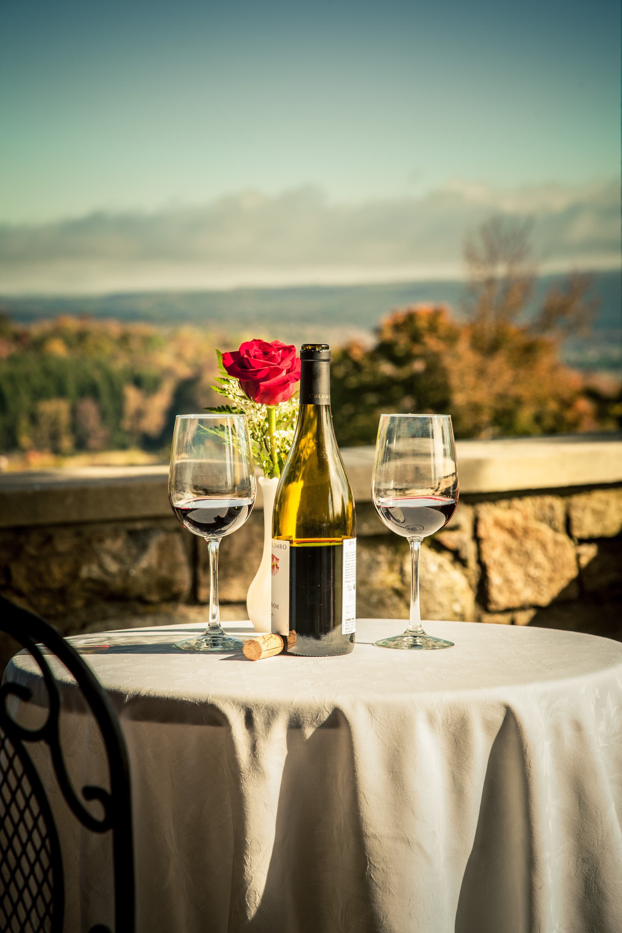 A table on a patio with a white tablecloth, a bottle of wine, two glasses of wine, and a bouquet of red roses. The background shows a scenic view of mountains and fall foliage.