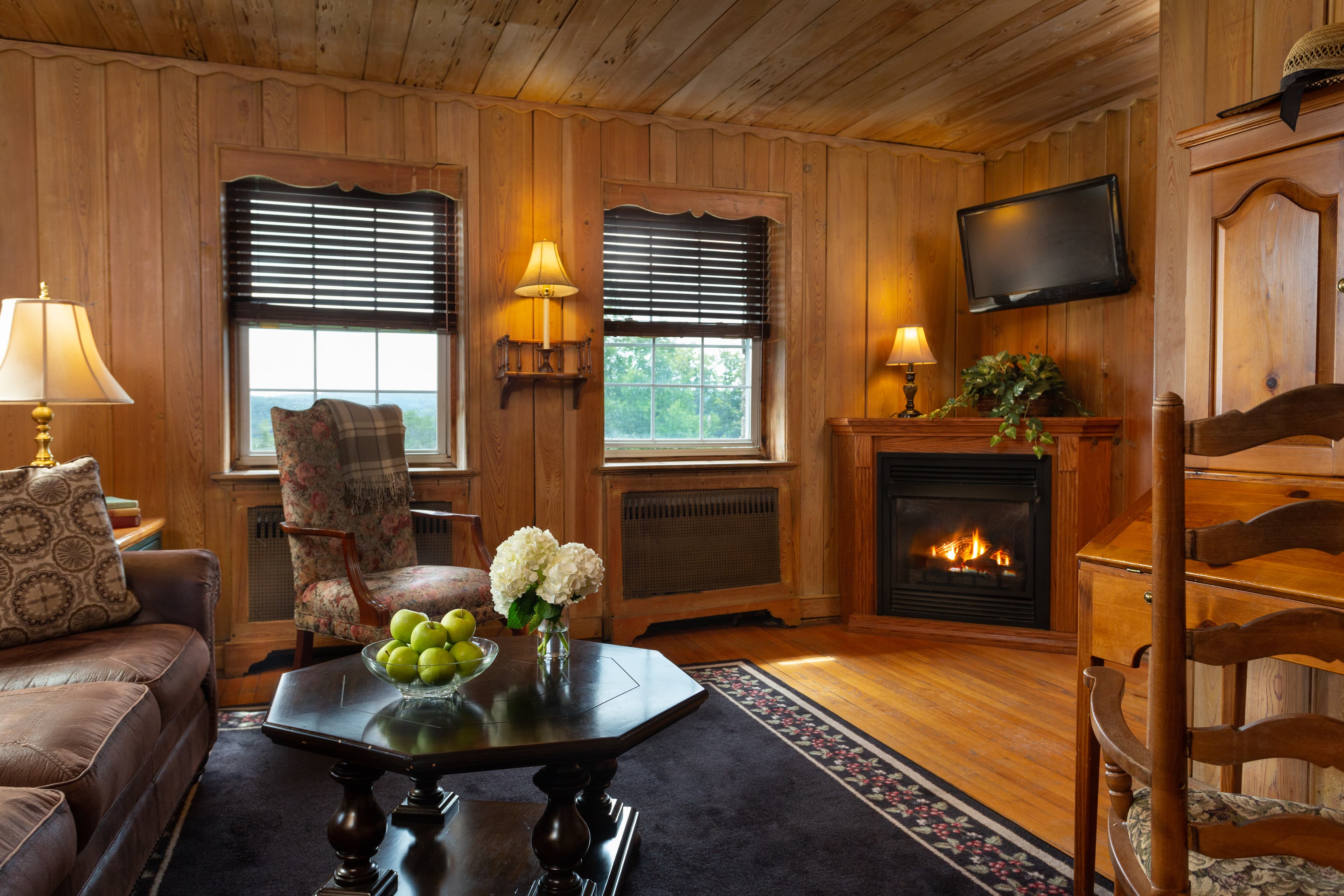 Cozy wooden living room with a fireplace, two windows, and a table of green apples.