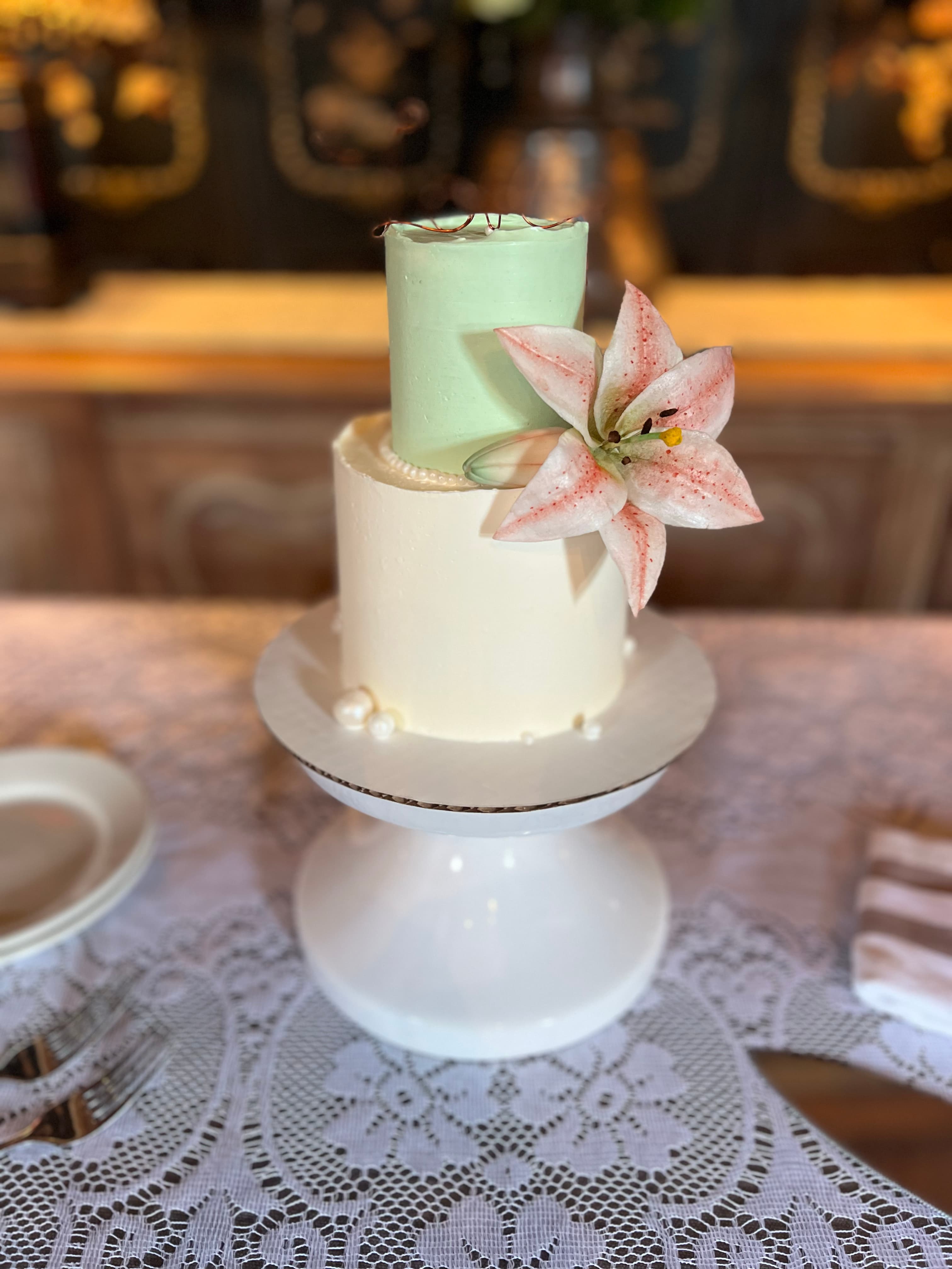 A two-tiered wedding cake with a green and white buttercream frosting, decorated with a pink lily flower.
