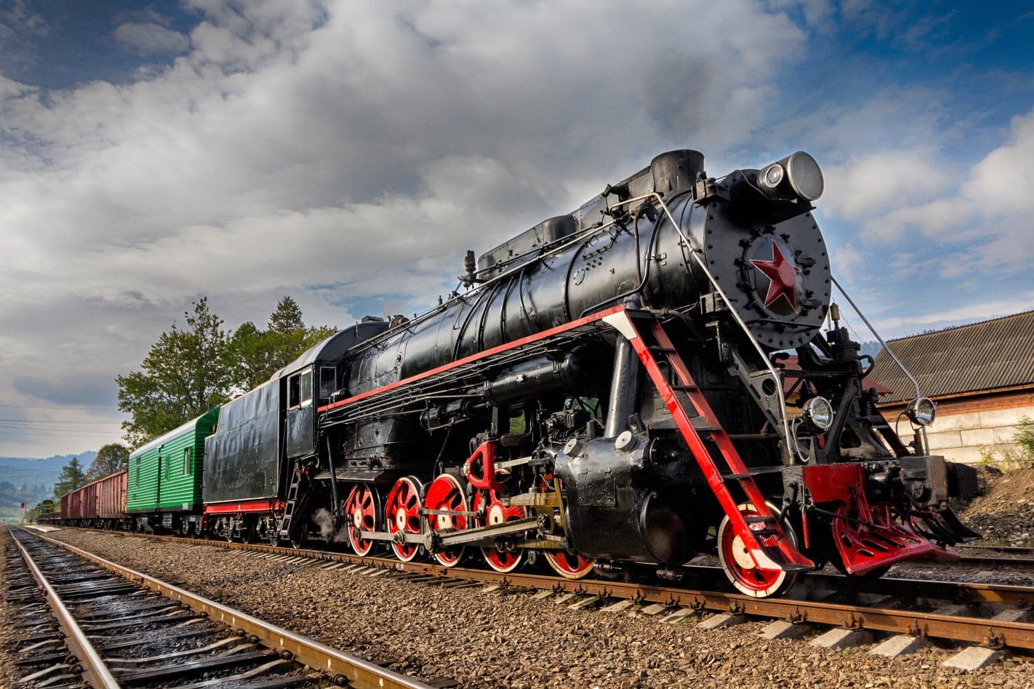 A black steam locomotive with red accents and green freight cars on a railway track under a cloudy sky.