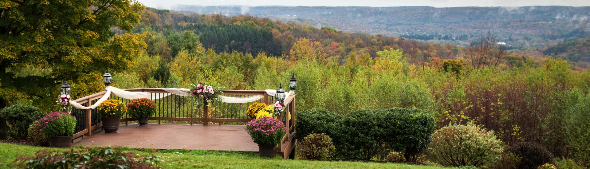 A scenic outdoor wedding venue with a wooden deck overlooking a valley. The deck is decorated with flowers and fabric, and there are lanterns on the sides. The valley below is filled with colorful fall foliage.