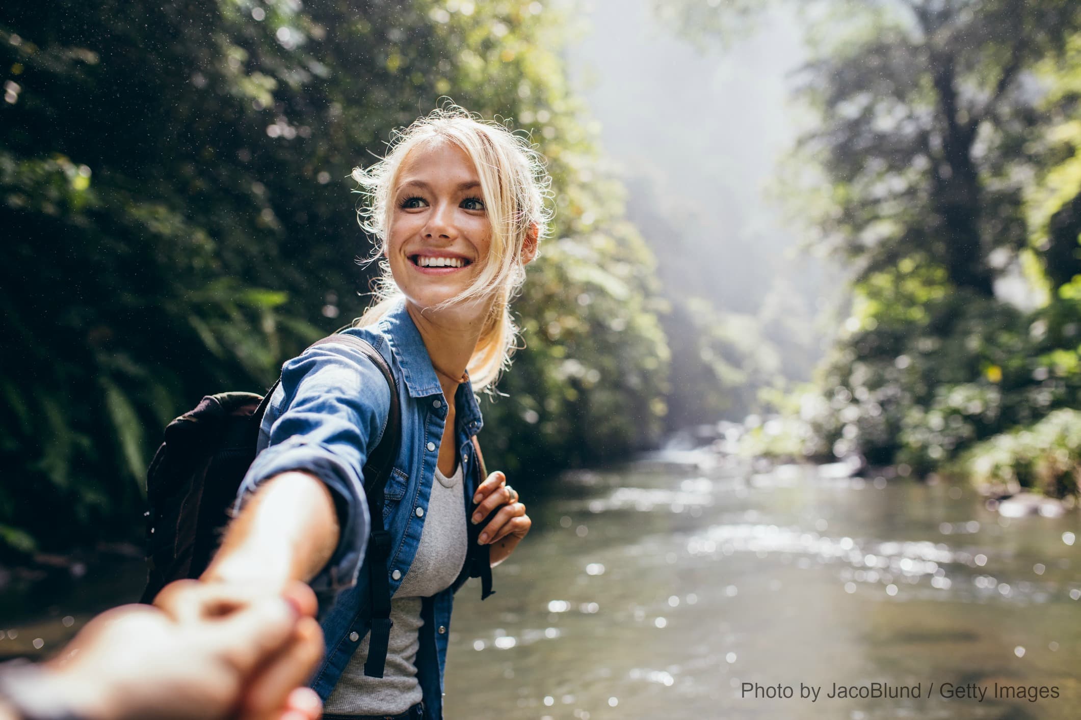 A smiling woman reaches out to hold hands while standing near a serene river surrounded by lush greenery. A smiling woman reaches out to hold hands while standing near a serene river surrounded by lush greenery.