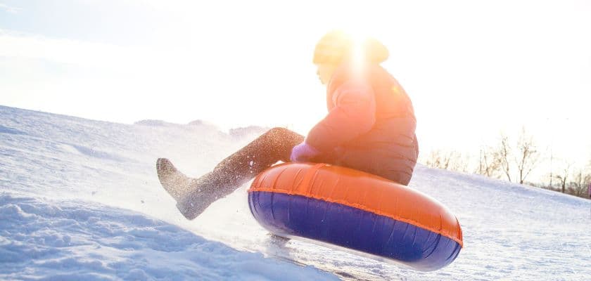 A person is sledding down a snowy hill on an orange and blue inflatable tube, with the sun shining brightly in the background.