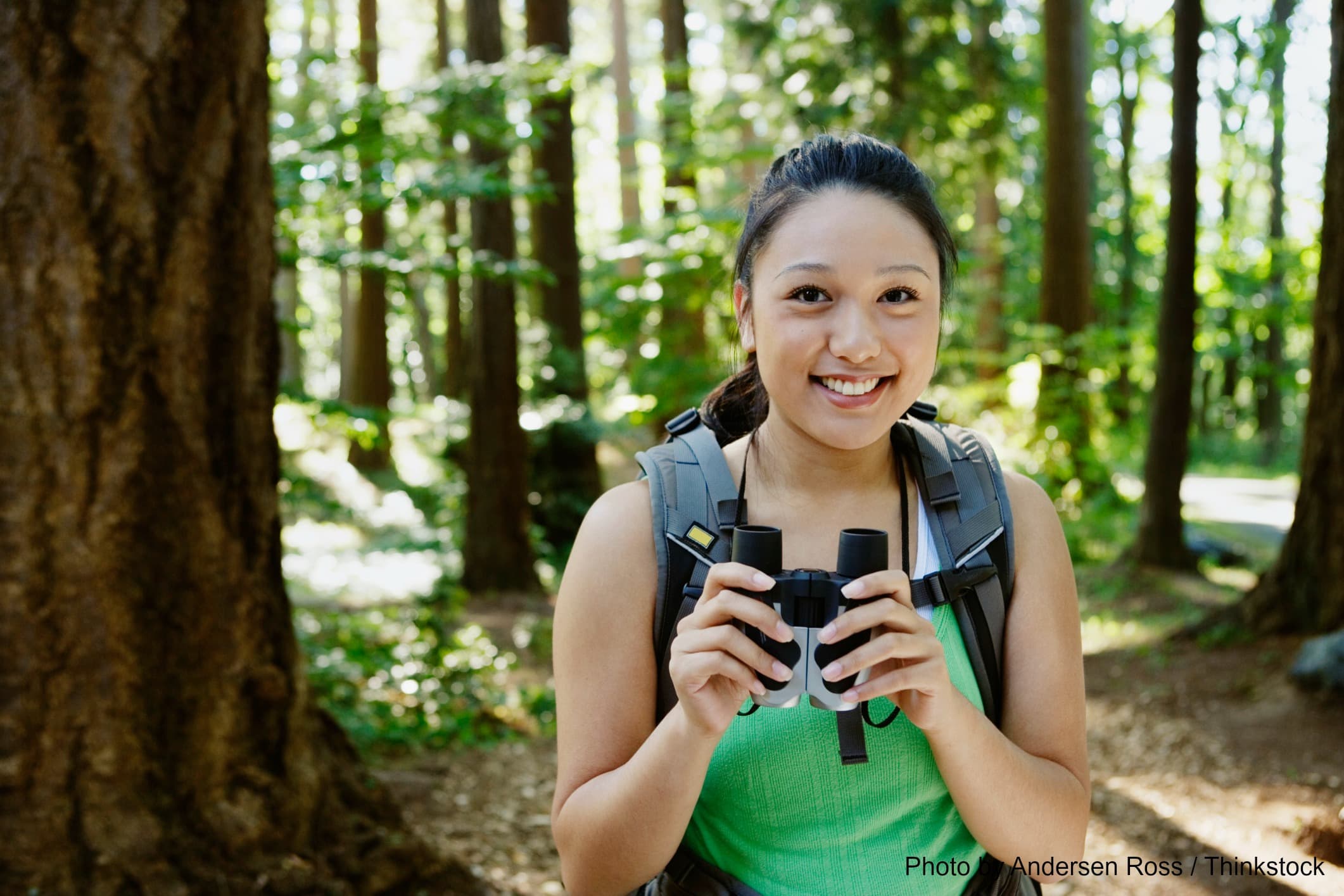 A smiling young woman holds binoculars while standing in a forest. A smiling young woman holds binoculars while standing in a forest.