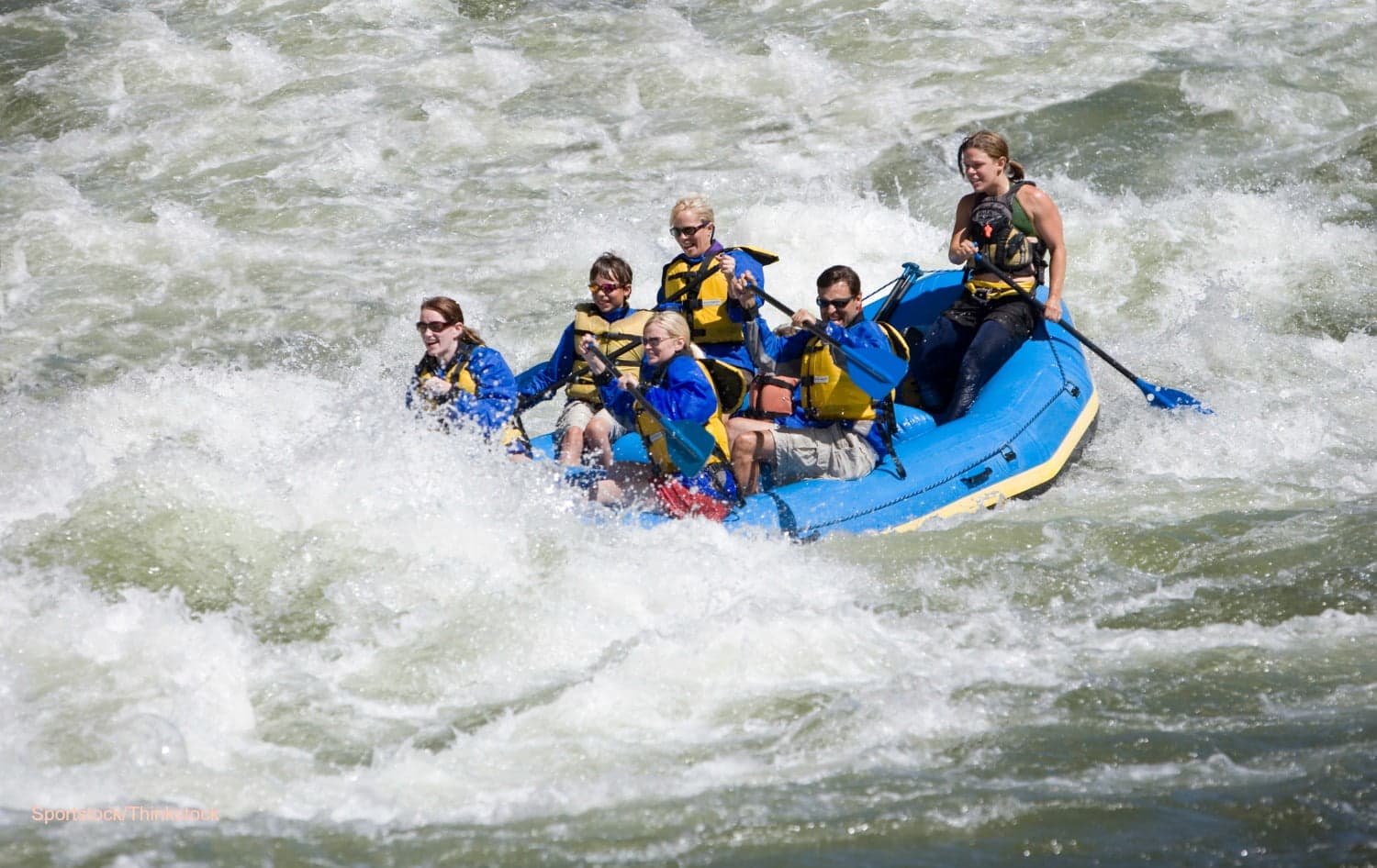 A group of six people paddles a blue raft through white water rapids.
