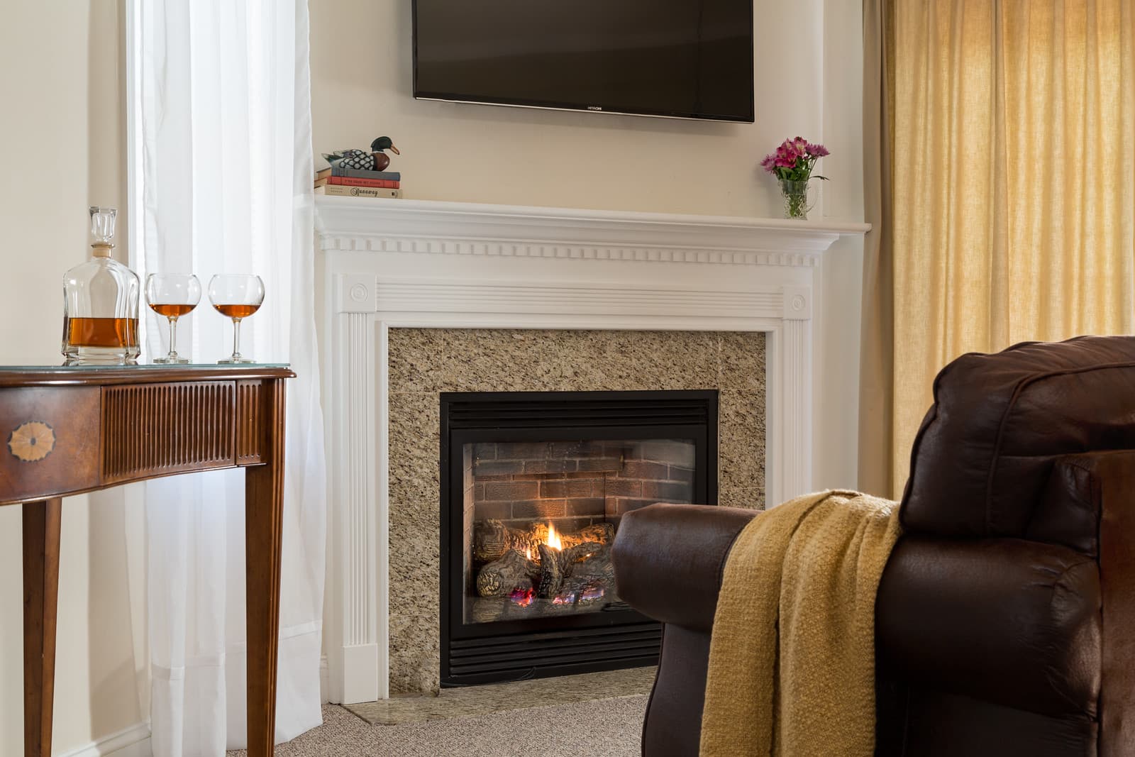 A cozy living room featuring a fireplace, a brown leather chair, a wooden side table with drinks, and a television mounted on the wall.
