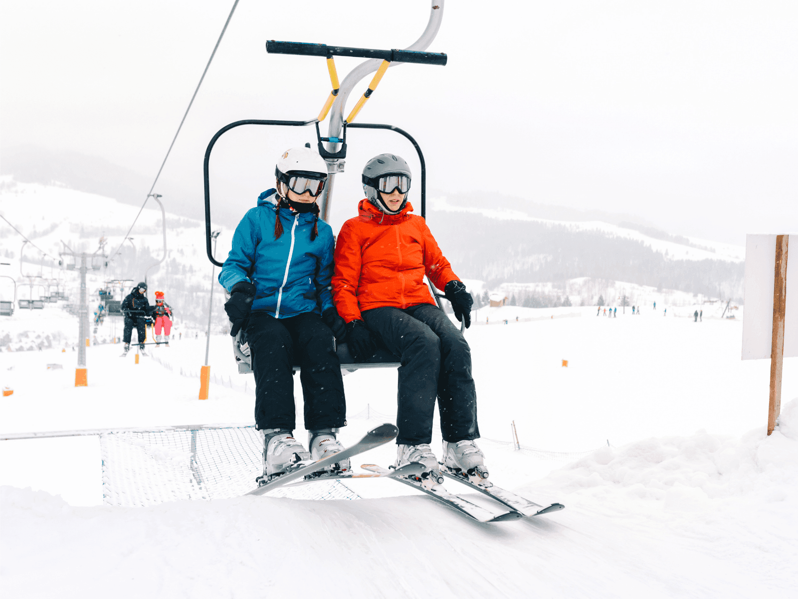 Two skiers wearing helmets and colorful jackets ride a ski lift in a snowy landscape. Two skiers wearing helmets and colorful jackets ride a ski lift in a snowy landscape.