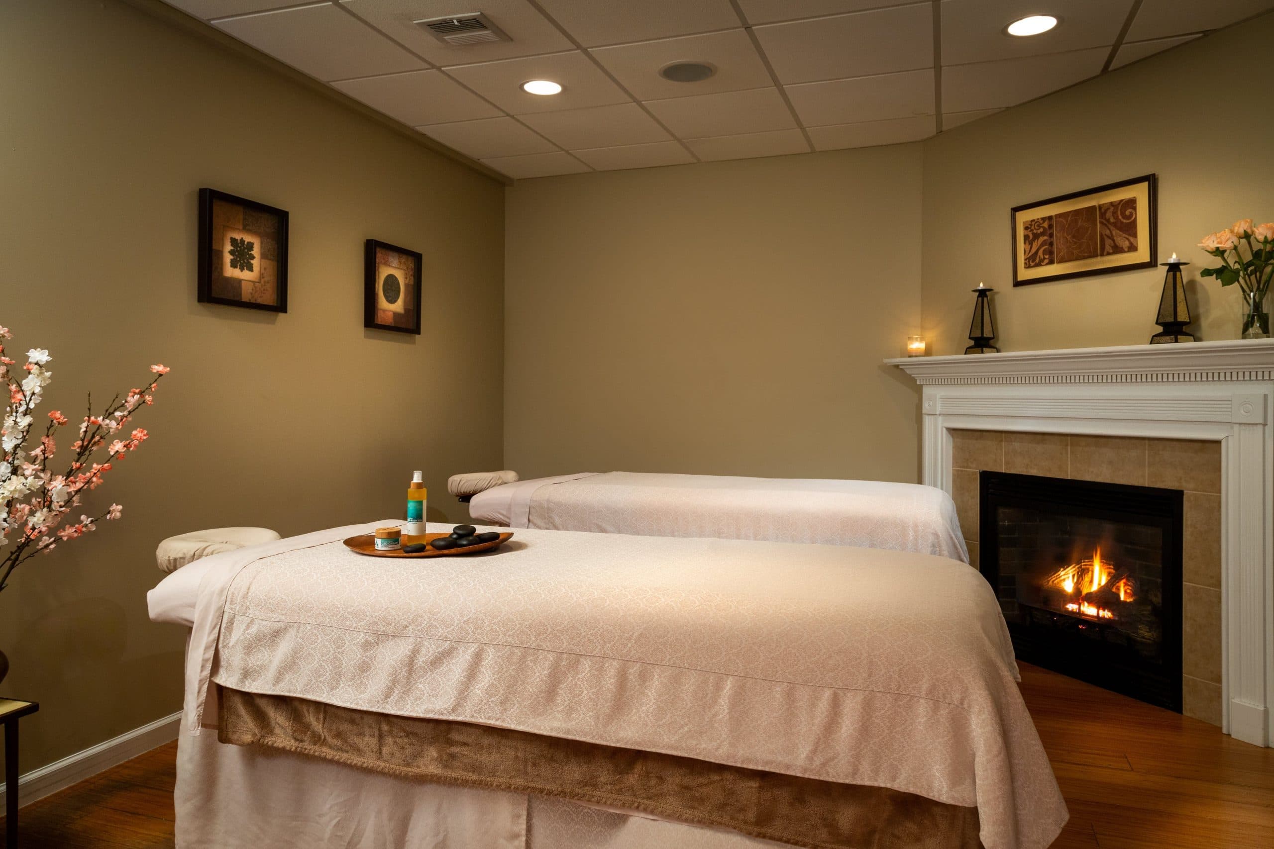 A relaxing spa room with two massage tables, each covered with white linens and towels. There is a fireplace with a crackling fire in the background, and the walls are decorated with artwork and a vase of flowers.