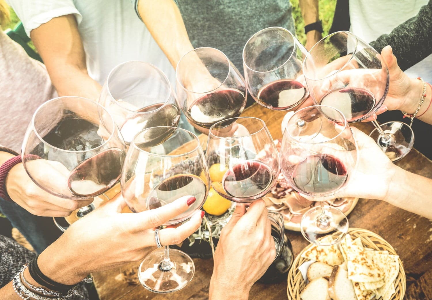 A group cheers with glasses of red wine around a wooden table with snacks.